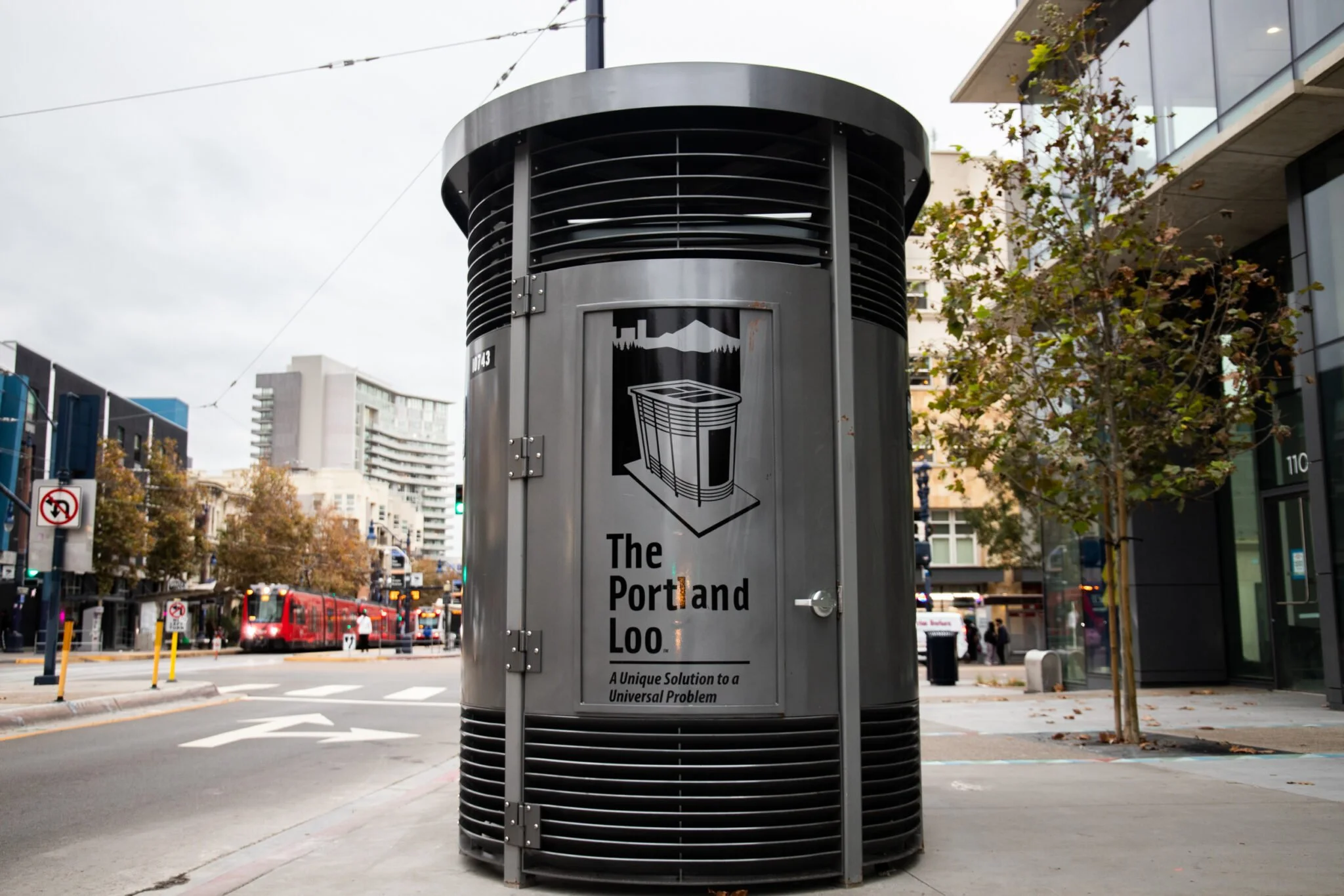 A cylindrical street furniture with a sign indicating it's part of the Portland Loo series, featuring a simple graphic of a portable toilet and text that reads "The Portland Loo" with a tagline about a unique solution.
