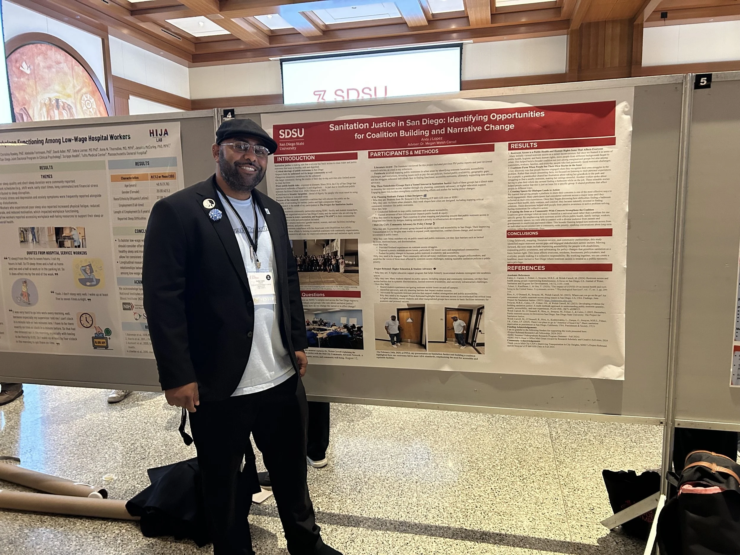 A man wearing a black blazer, white t-shirt, glasses, and a black cap smiling at the camera. He is standing in front of research posters displayed on boards in an indoor conference or academic event setting.