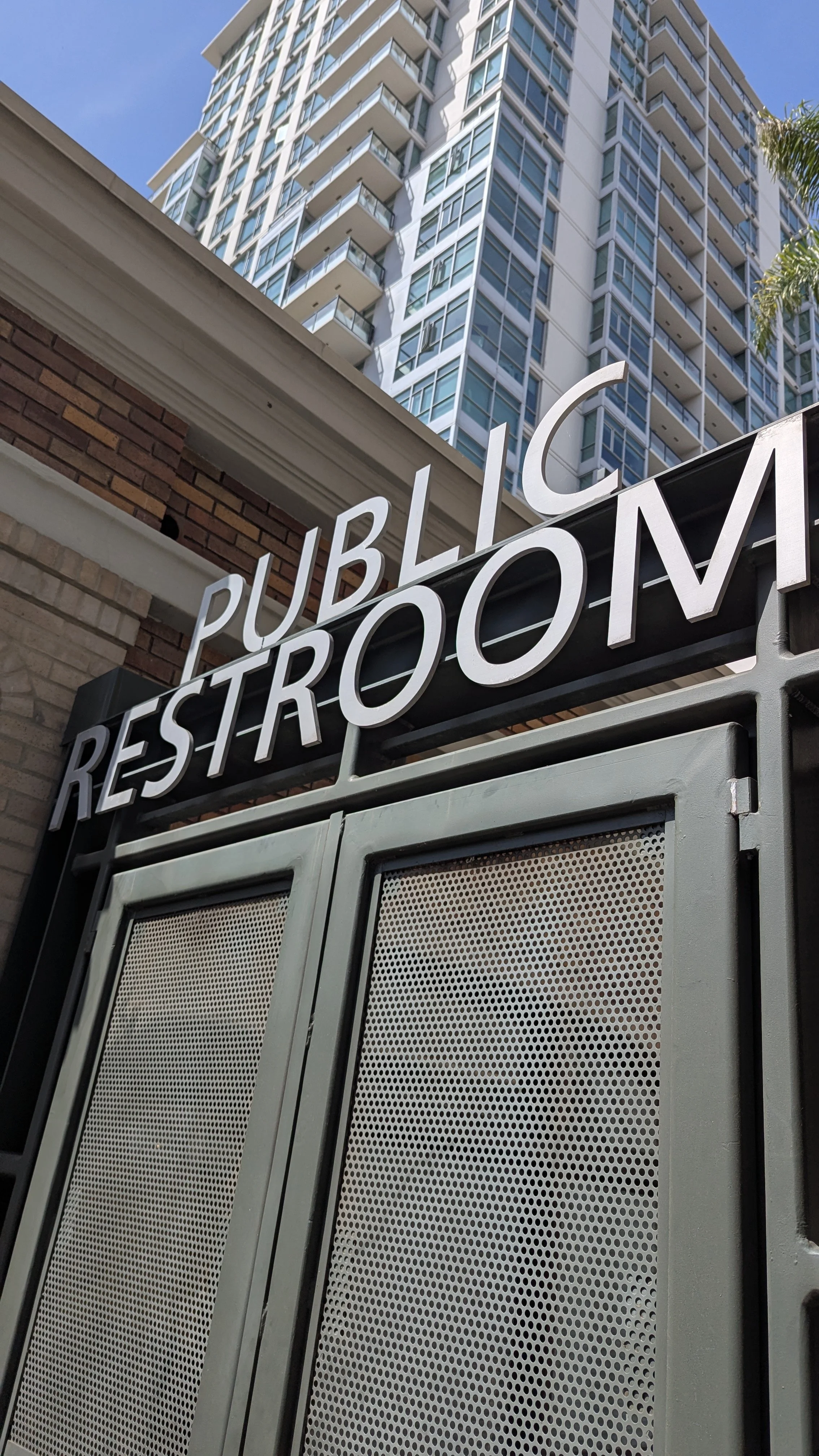 Close-up of a metal restaurant or pub sign reading 'PUBLIC RESTROOM' in front of a tall modern high-rise building with balconies, under a clear blue sky.