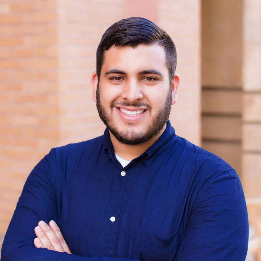 A smiling man with dark hair, beard, and fair skin, wearing a navy blue button-up shirt, standing outdoors in front of a brick wall.