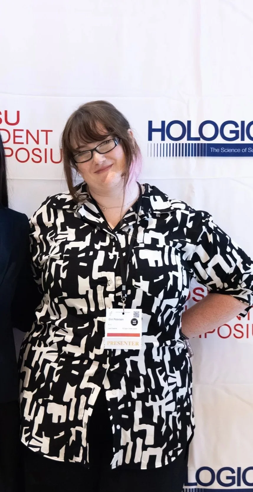 A woman with glasses and shoulder-length brown hair, wearing a black and white patterned blouse, standing in front of a backdrop with the words "Hologic" and "Student Symposium," smiling with her eyes closed.