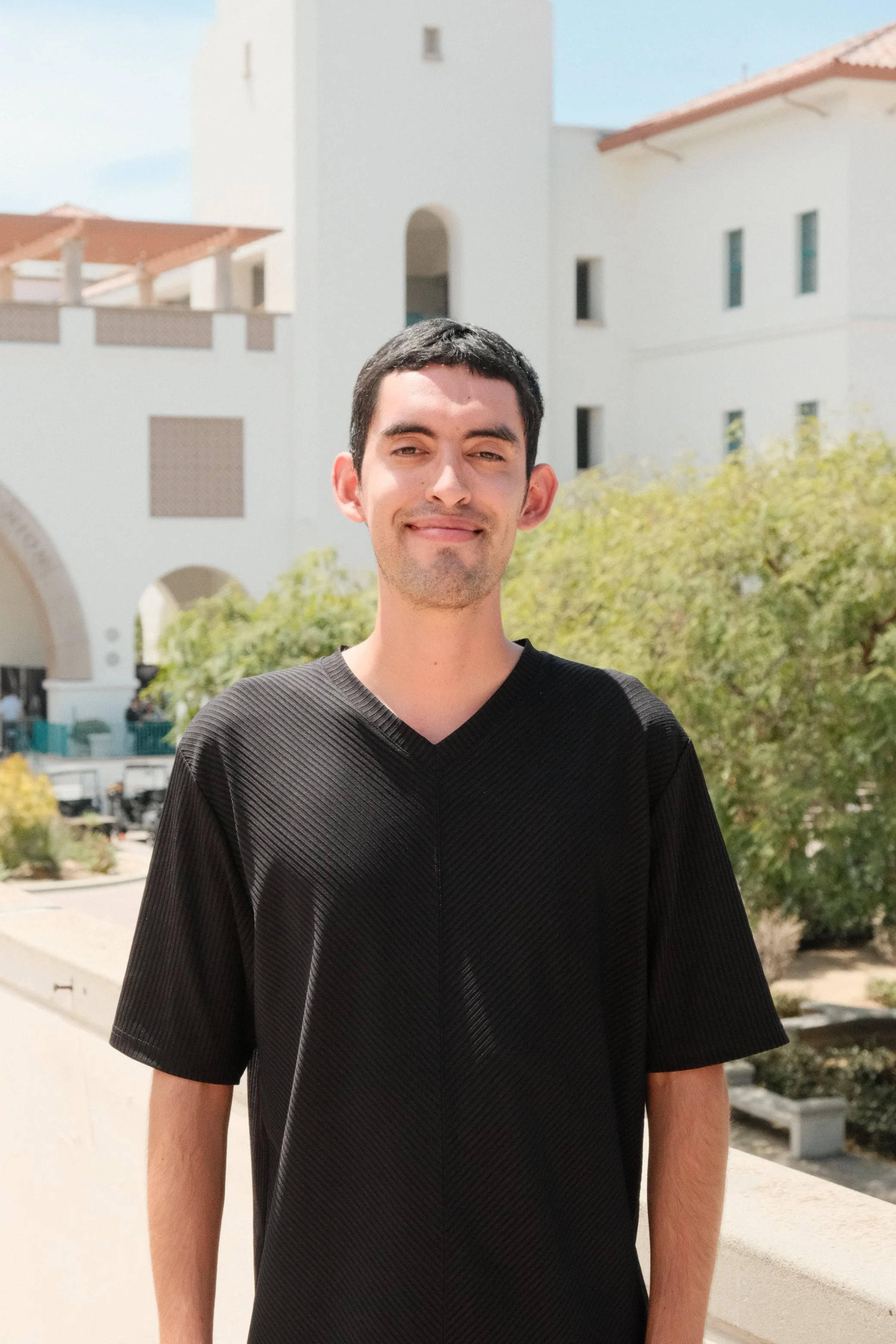A young man with black hair, wearing a black shirt, standing outside in front of a beige building with trees and bushes in the background.
