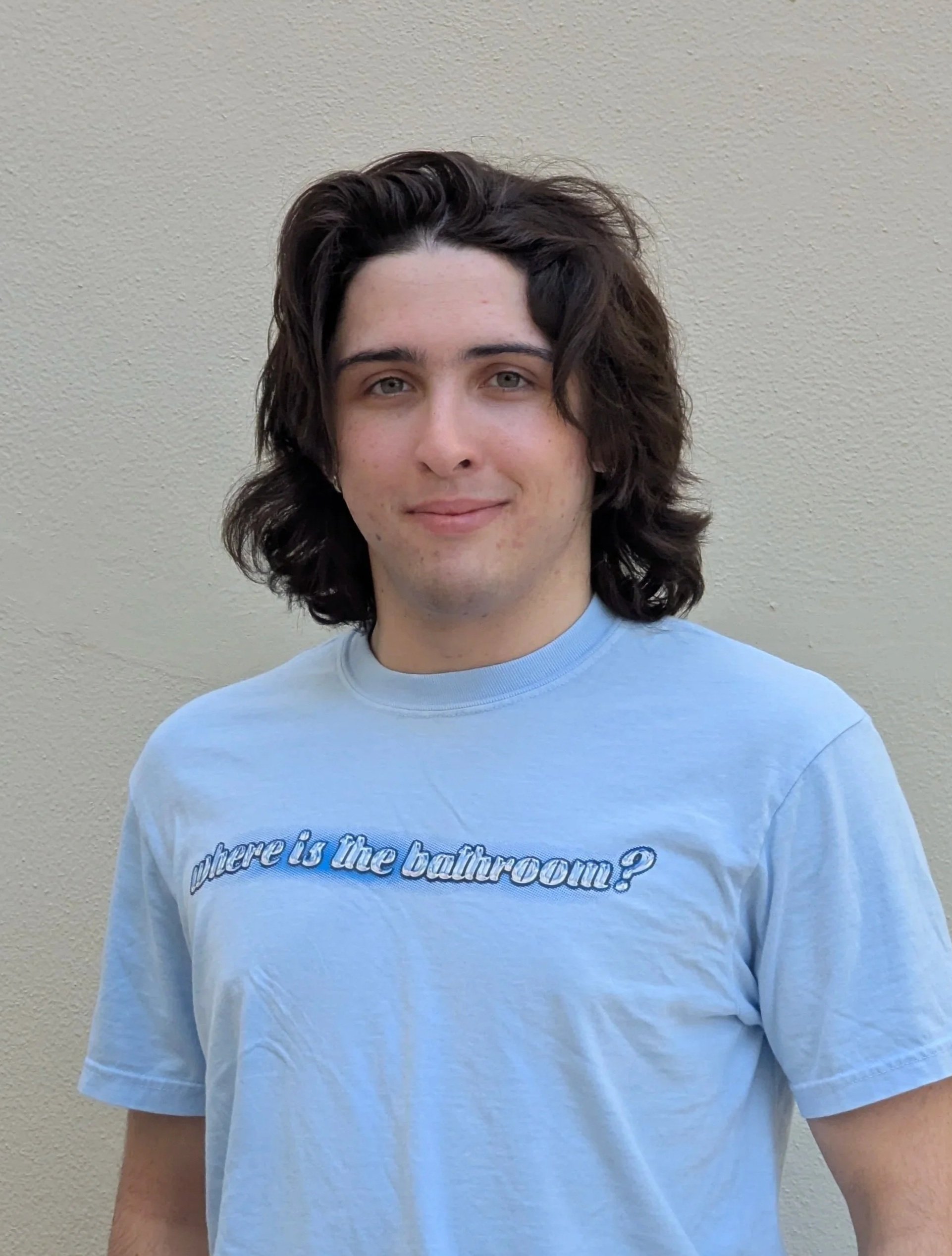 Young person with long, wavy dark hair, wearing a light blue T-shirt with text, standing against a beige wall.