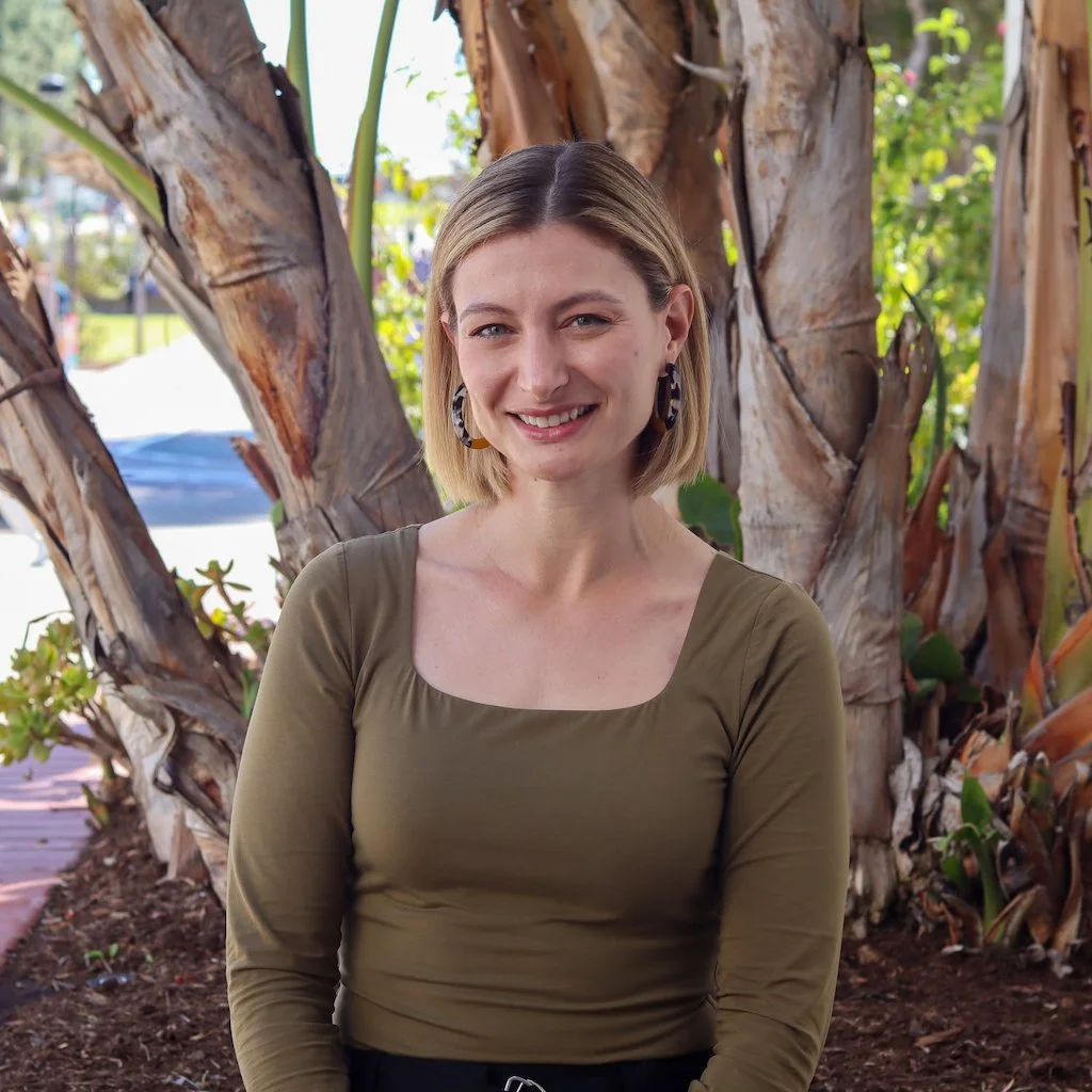 A young woman with short blonde hair, wearing a green long-sleeve top and large earrings, smiling outdoors with trees and greenery in the background.