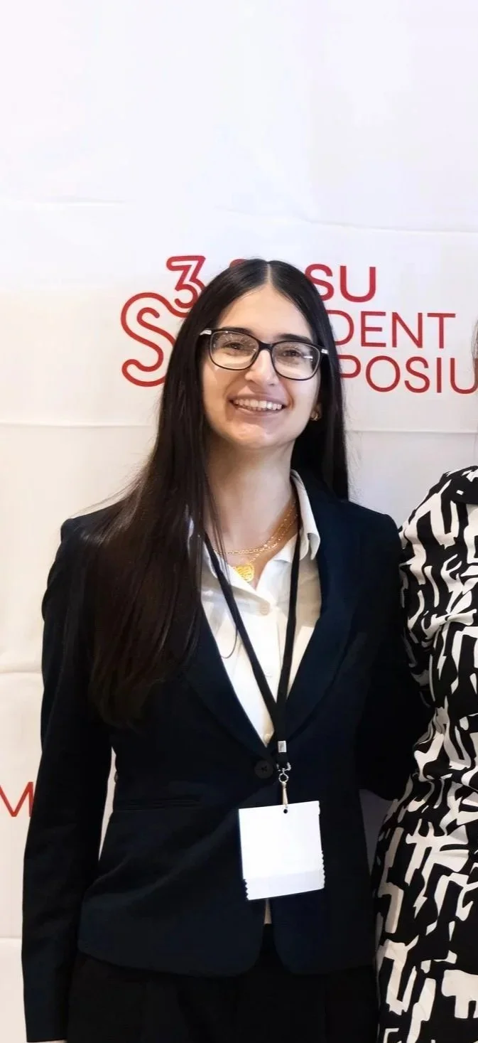 Young woman with glasses, long dark hair, wearing a black blazer, white shirt, and a lanyard, smiling in front of a white background with red text.
