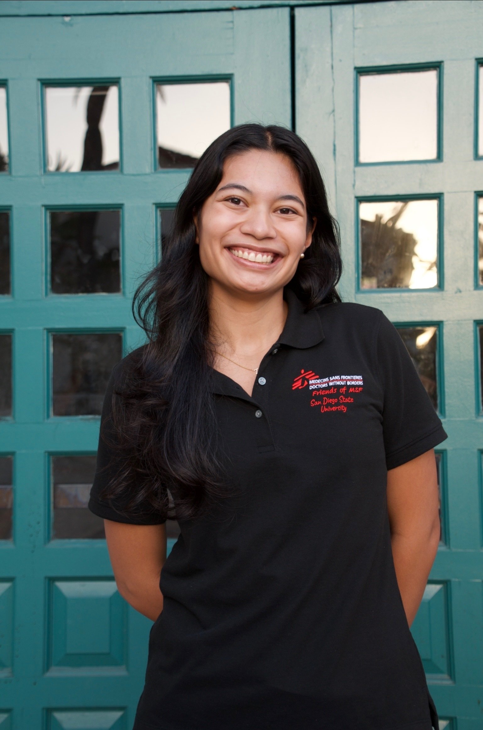 A young woman with long black hair, smiling and wearing a black polo shirt with a logo that says Friends of MSF, standing in front of a teal green window/door.