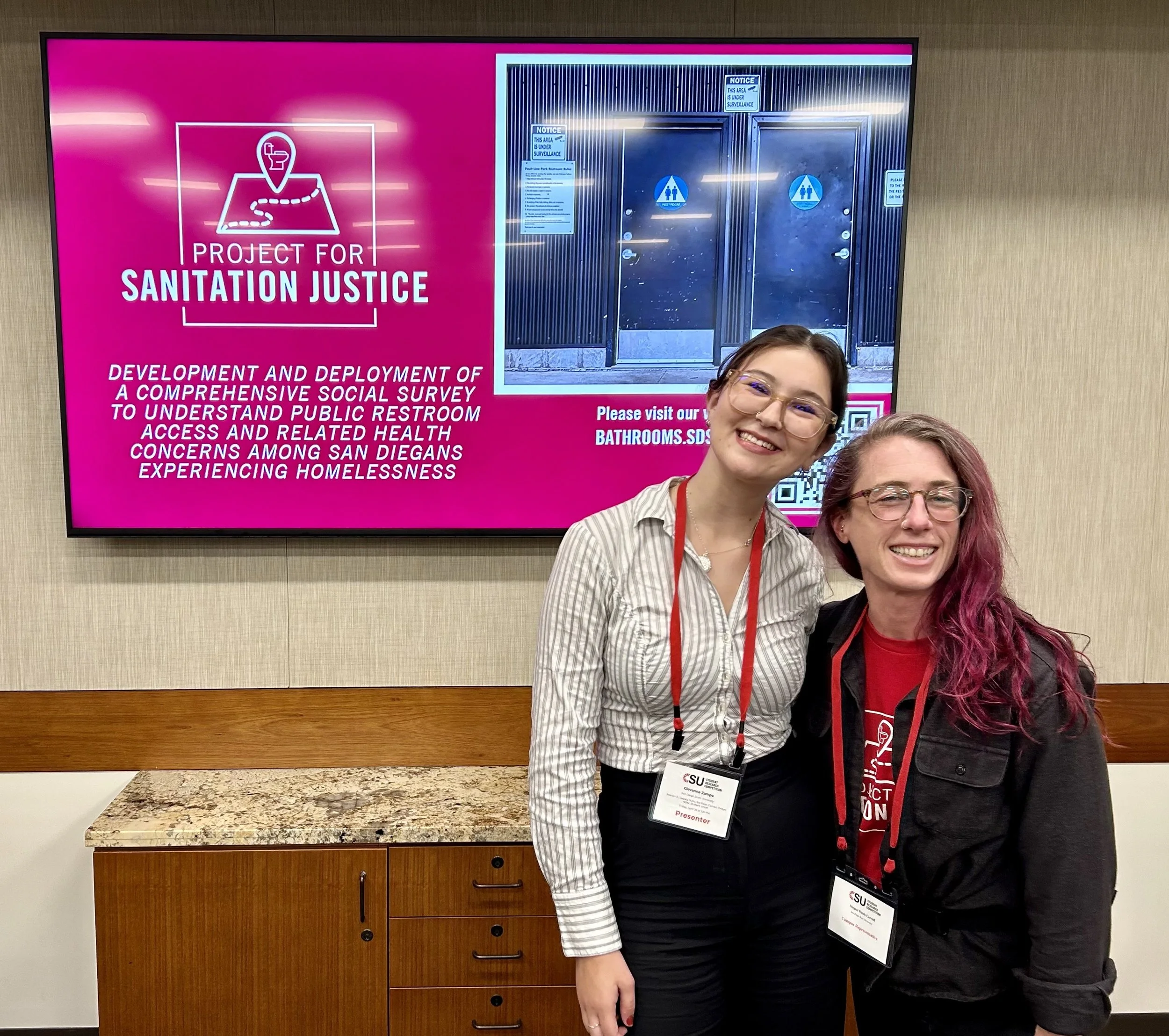 Two women smiling standing in front of a large presentation screen that reads "Project for Sanitation Justice" with additional text about developing and deploying a social survey related to public restroom access and health, with a wooden countertop and beige wall behind them.