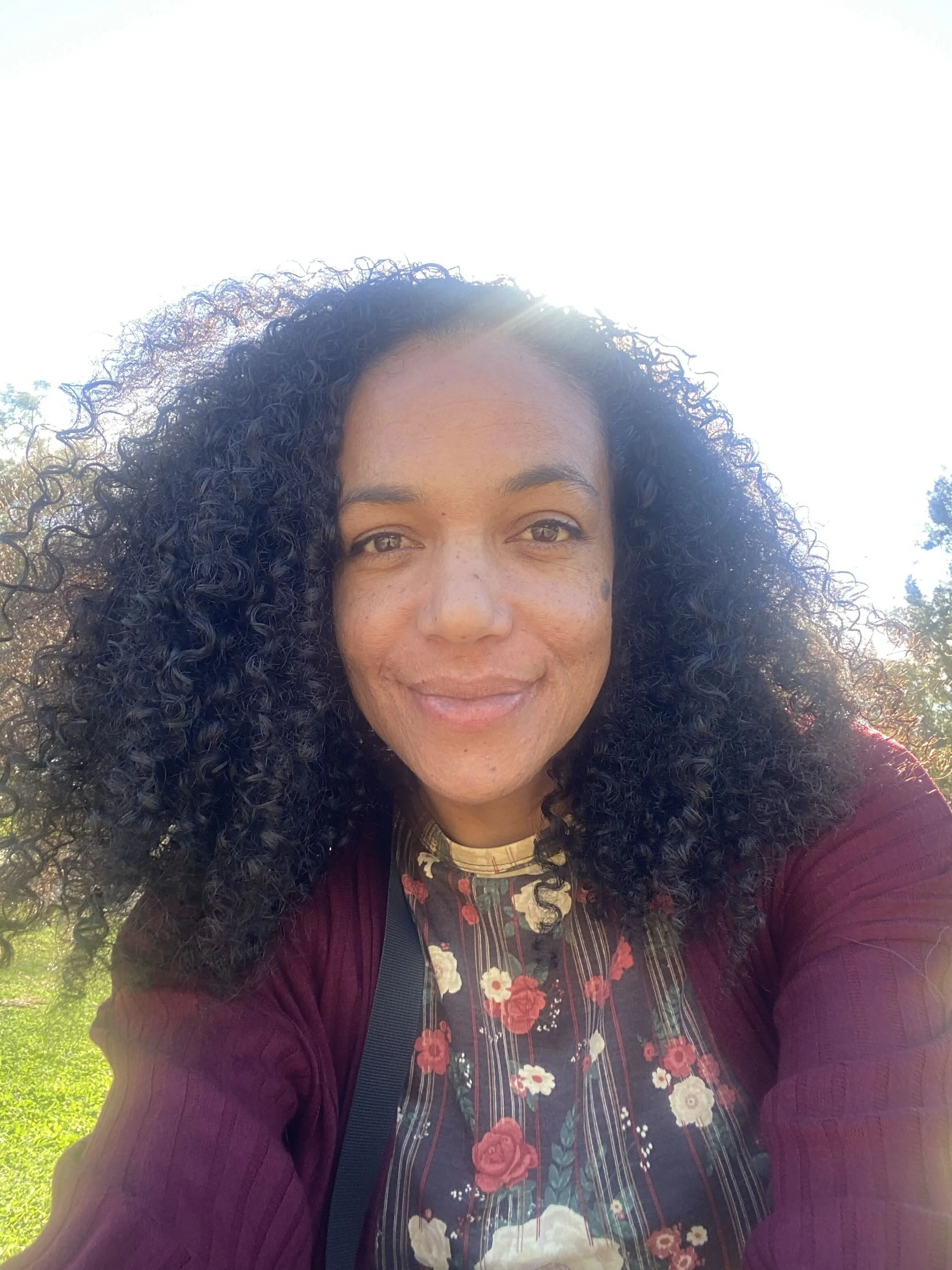 A woman with curly black hair taking a selfie outdoors with bright sunlight, grass, and trees in the background.