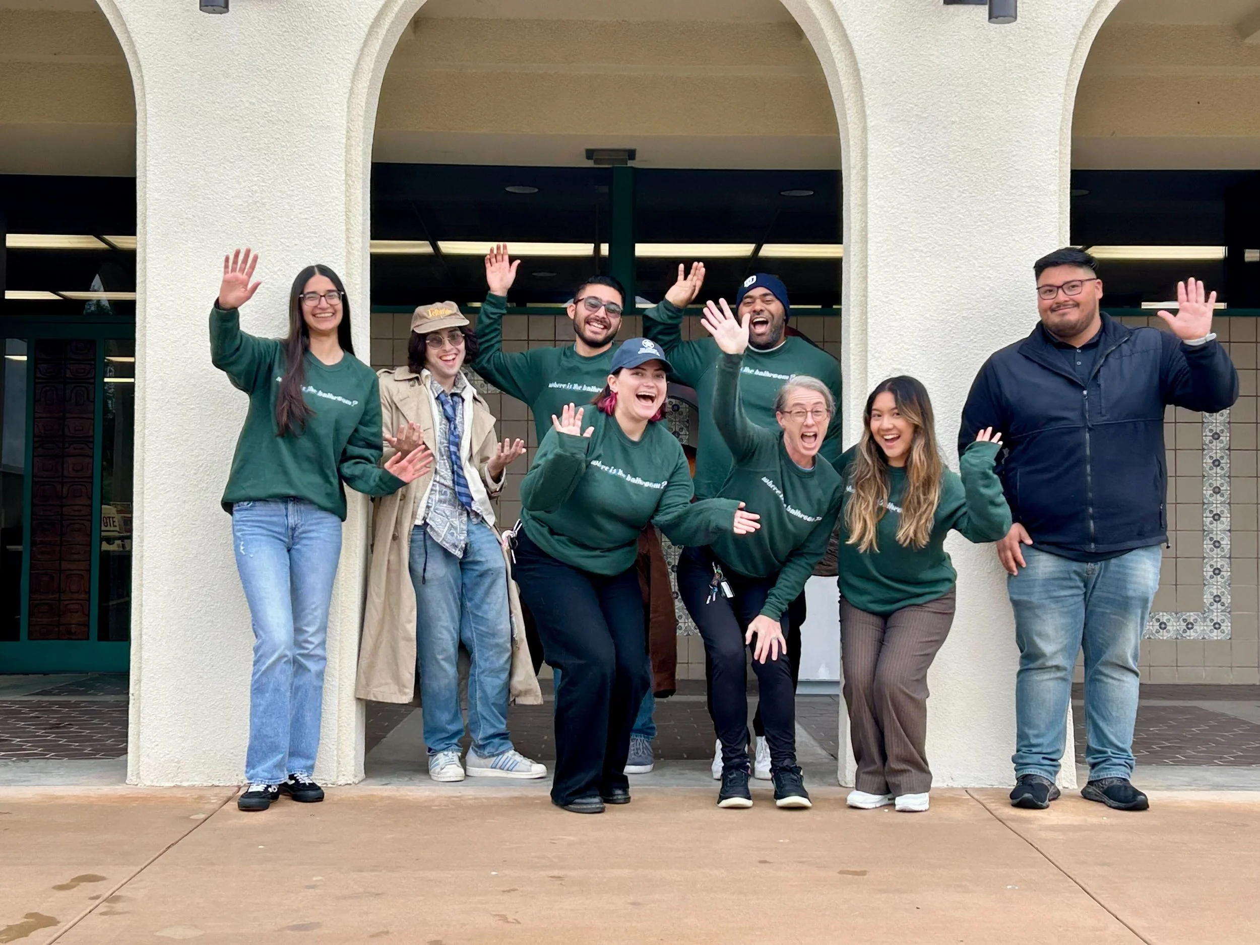 Group of diverse people standing in front of a building, smiling, waving, and enjoying each other's company.