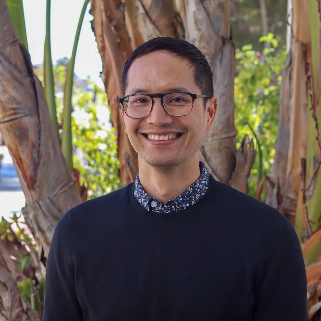 A young man with glasses and short dark hair smiling outdoors, standing in front of large banana plants with green leaves and brown trunks.