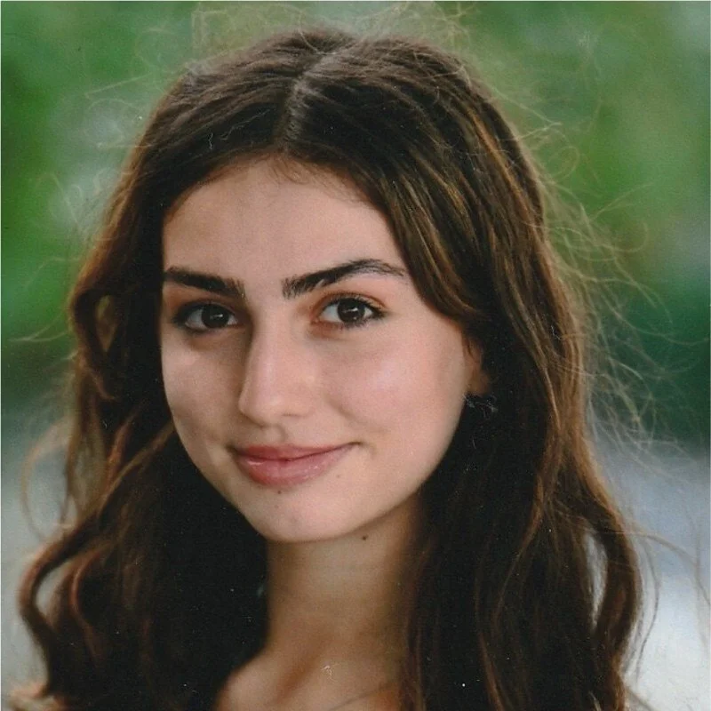 Close-up portrait of a young woman with wavy brown hair and light skin, smiling softly outdoors.