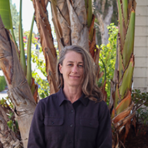 A woman with long, wavy gray hair standing outdoors in front of a large tree with broad leaves, wearing a dark button-up shirt.