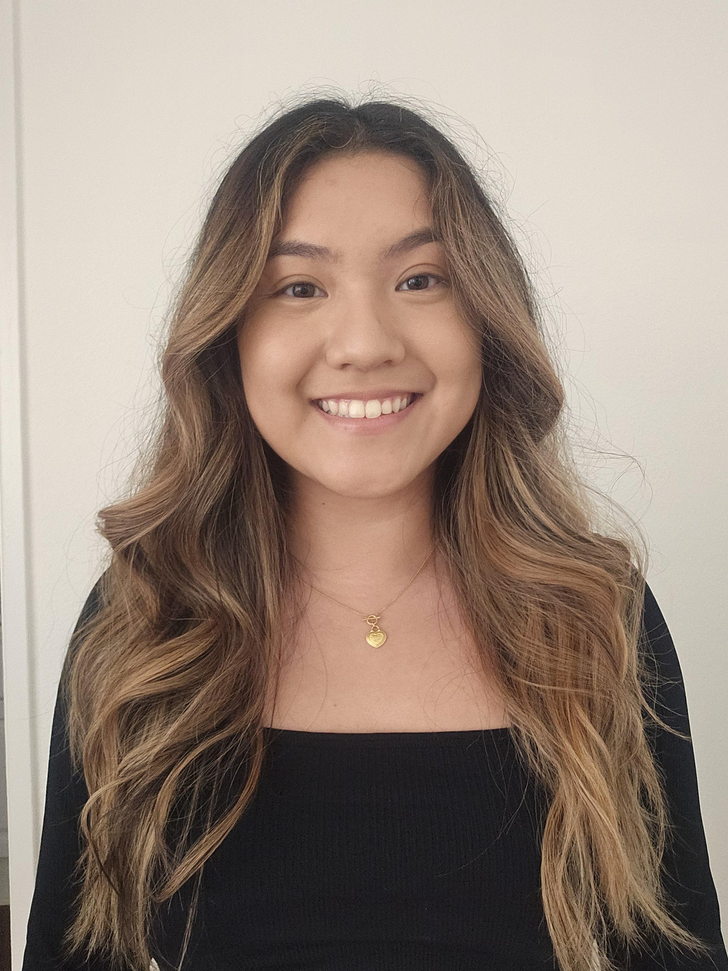 Close-up of a smiling woman with long, wavy hair with highlights, wearing a black top and a gold heart necklace, standing against a plain white background.