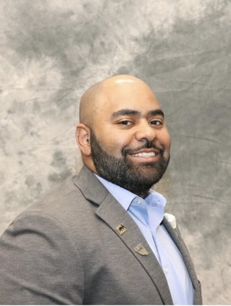 A smiling man with a dark beard and a shaved head, wearing a gray suit jacket and light blue shirt, posing against a cloudy gray background.