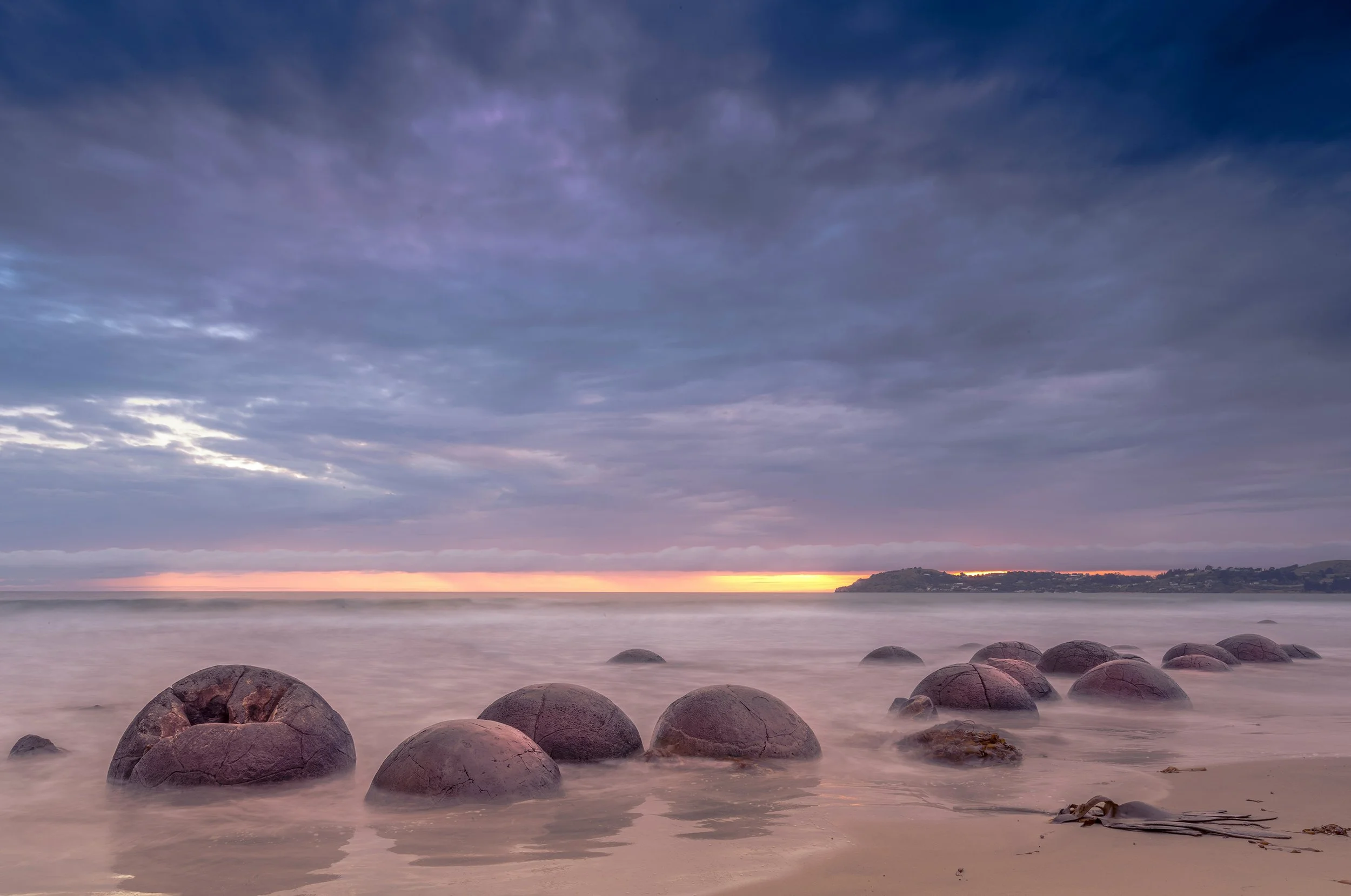 Photo of a calm ocean with large rocks in the foreground, a distant shoreline, and a cloudy sky during sunset with pink and purple hues.