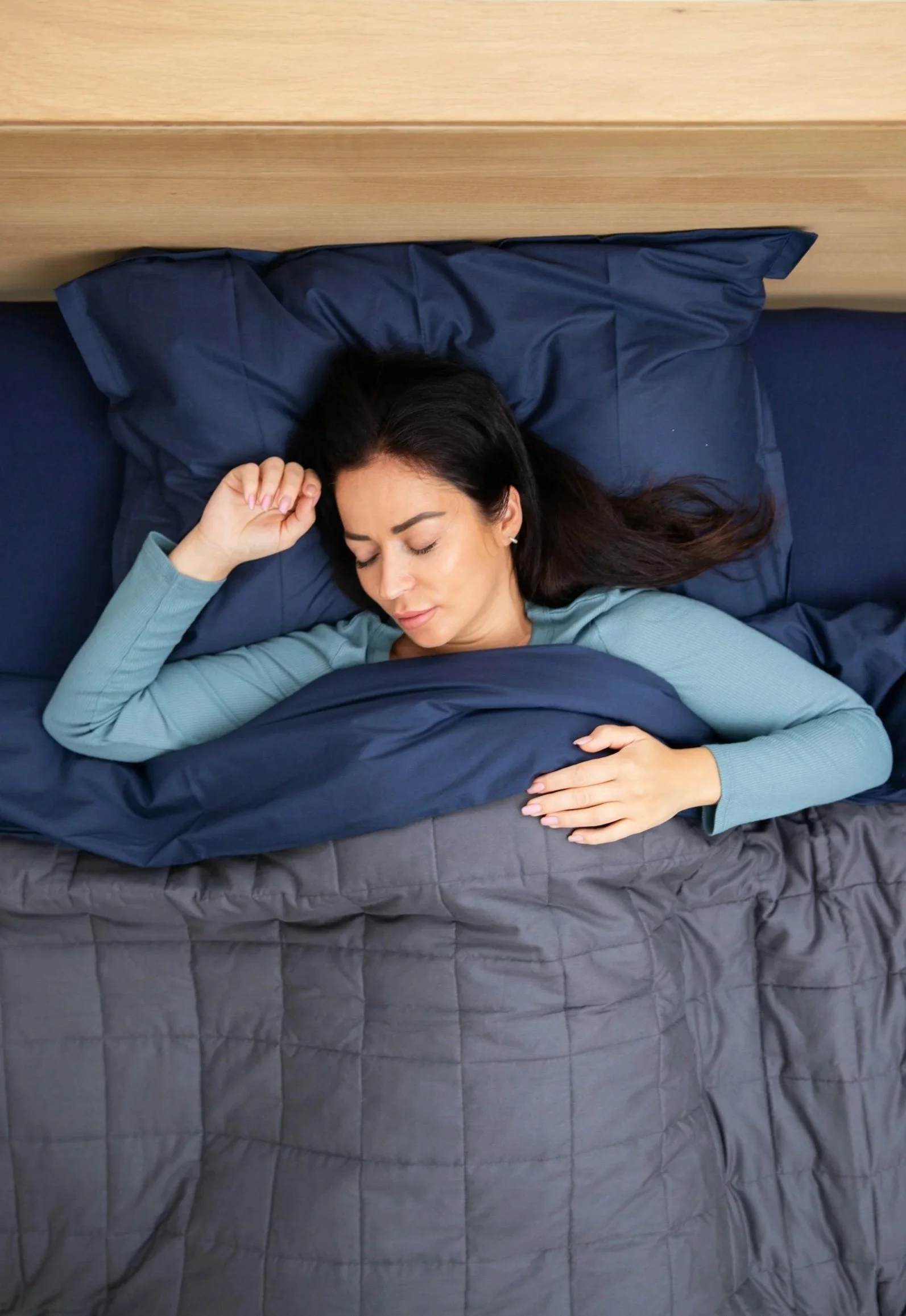 Woman with long dark hair sleeping on a bed with a dark gray quilt, pillow, and a blue pillowcase, covered in a light blue long-sleeved shirt, relaxing with eyes closed.