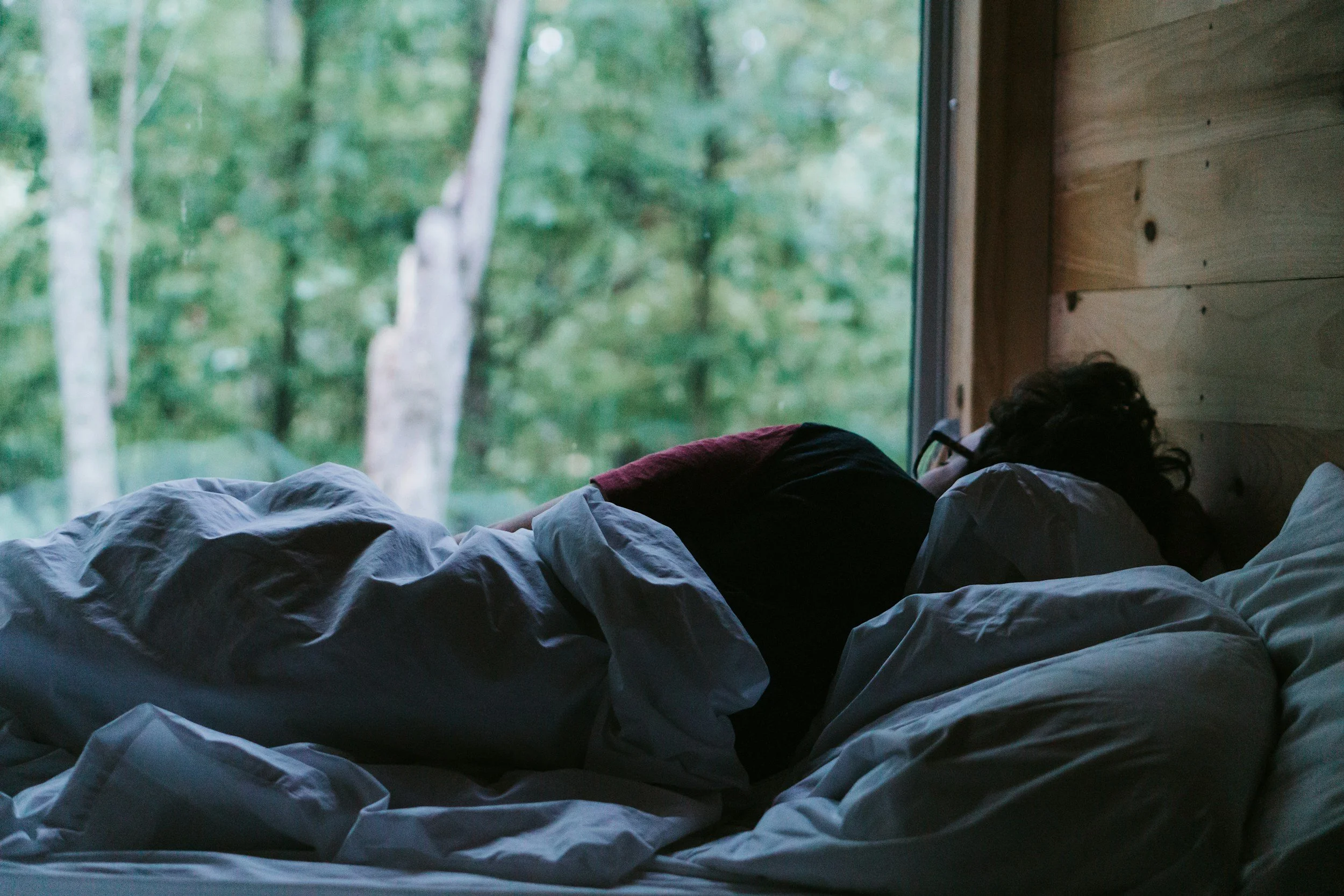 Person lying in bed with a view of green trees outside the window.