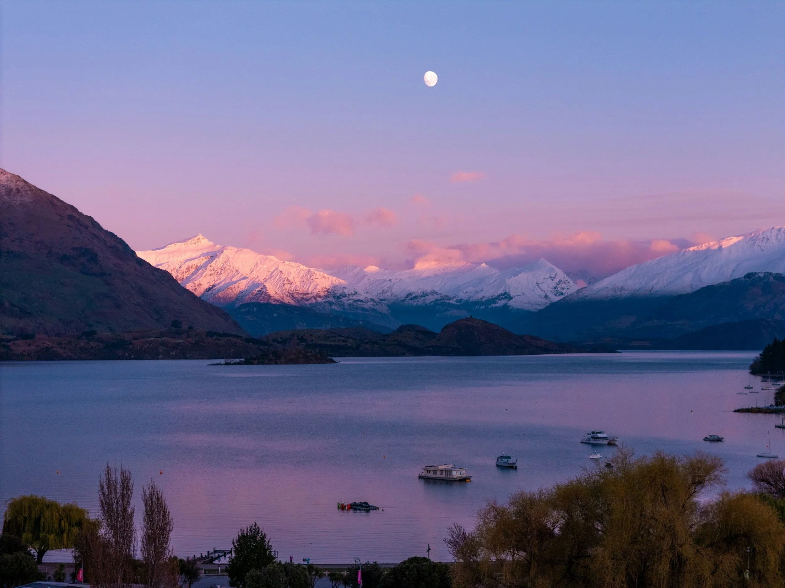 Scenic view of snow-capped mountains under a pastel twilight sky with the moon visible, overlooking a calm lake with boats and trees in the foreground.