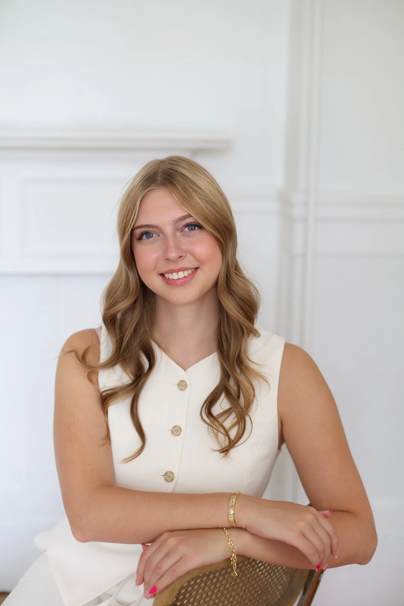 A young woman with wavy light brown hair and blue eyes smiling, sitting against a white background. She is wearing a sleeveless white button-up top and gold jewelry, including bracelets and rings.