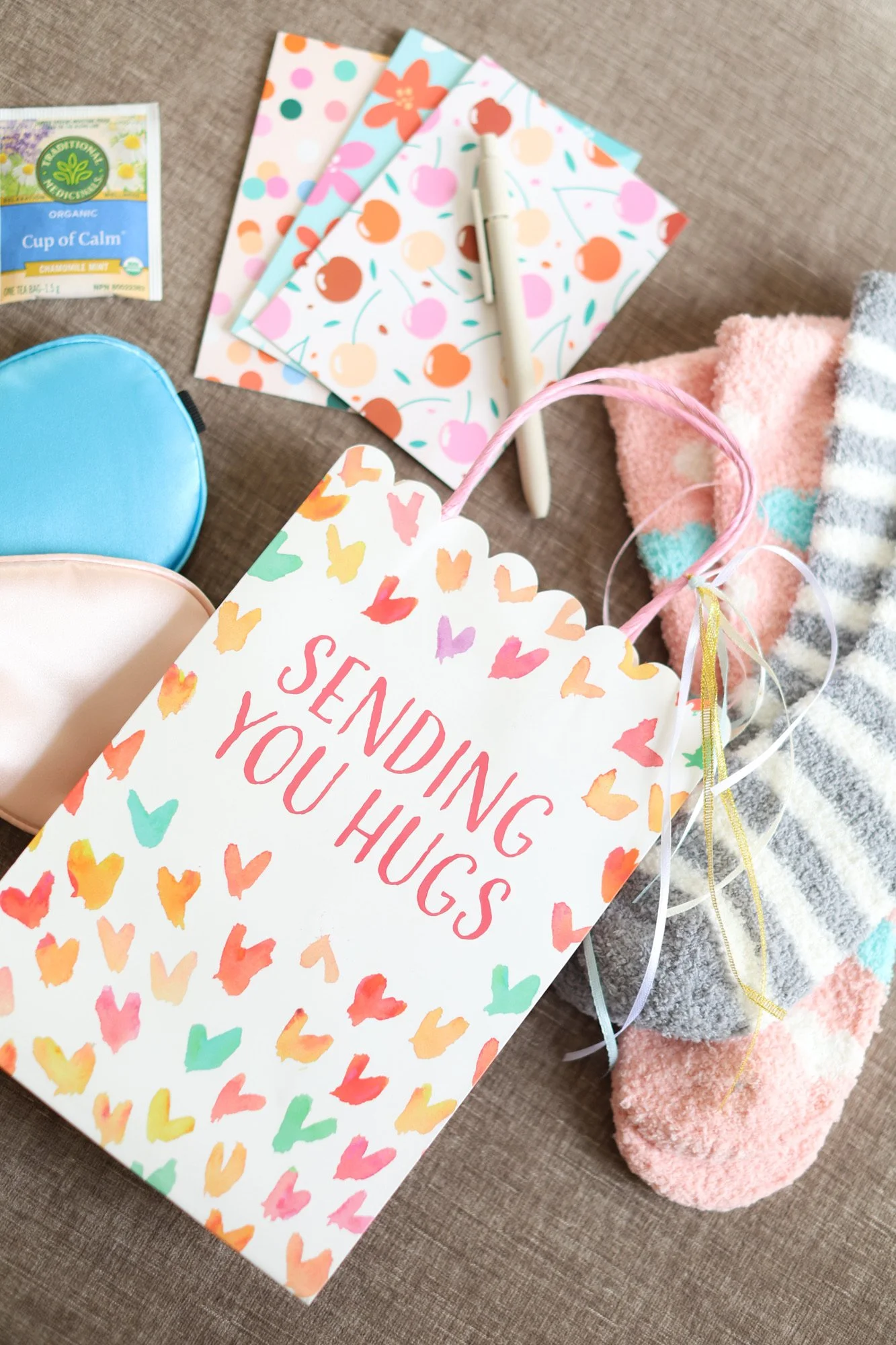 Colorful gift bag with hearts and the message "Sending you hugs" on it, along with a pink and gray striped sock, a pink and blue fuzzy item, a stack of three patterned notebooks, a pen, a tea bag packet labeled "Cup of Calm," and some ribbons.