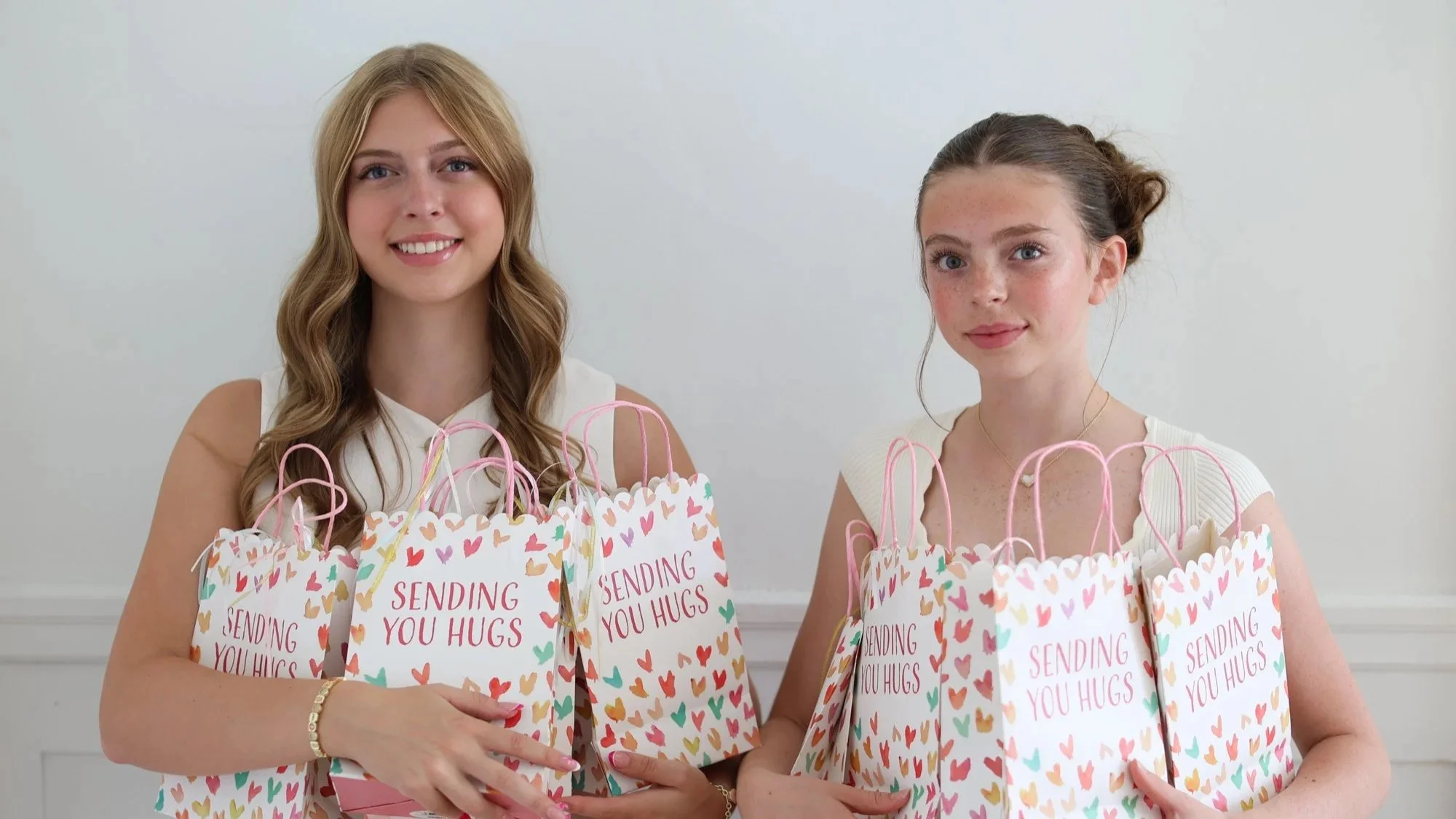 Two women standing side by side, each holding gift bags decorated with multicolored hearts and the words "Sending You Hugs." The woman on the left has long, wavy blonde hair and is smiling, wearing a white sleeveless top. The woman on the right has dark blonde or light brown hair tied back, with freckles, and is also smiling, wearing a white sleeveless top.