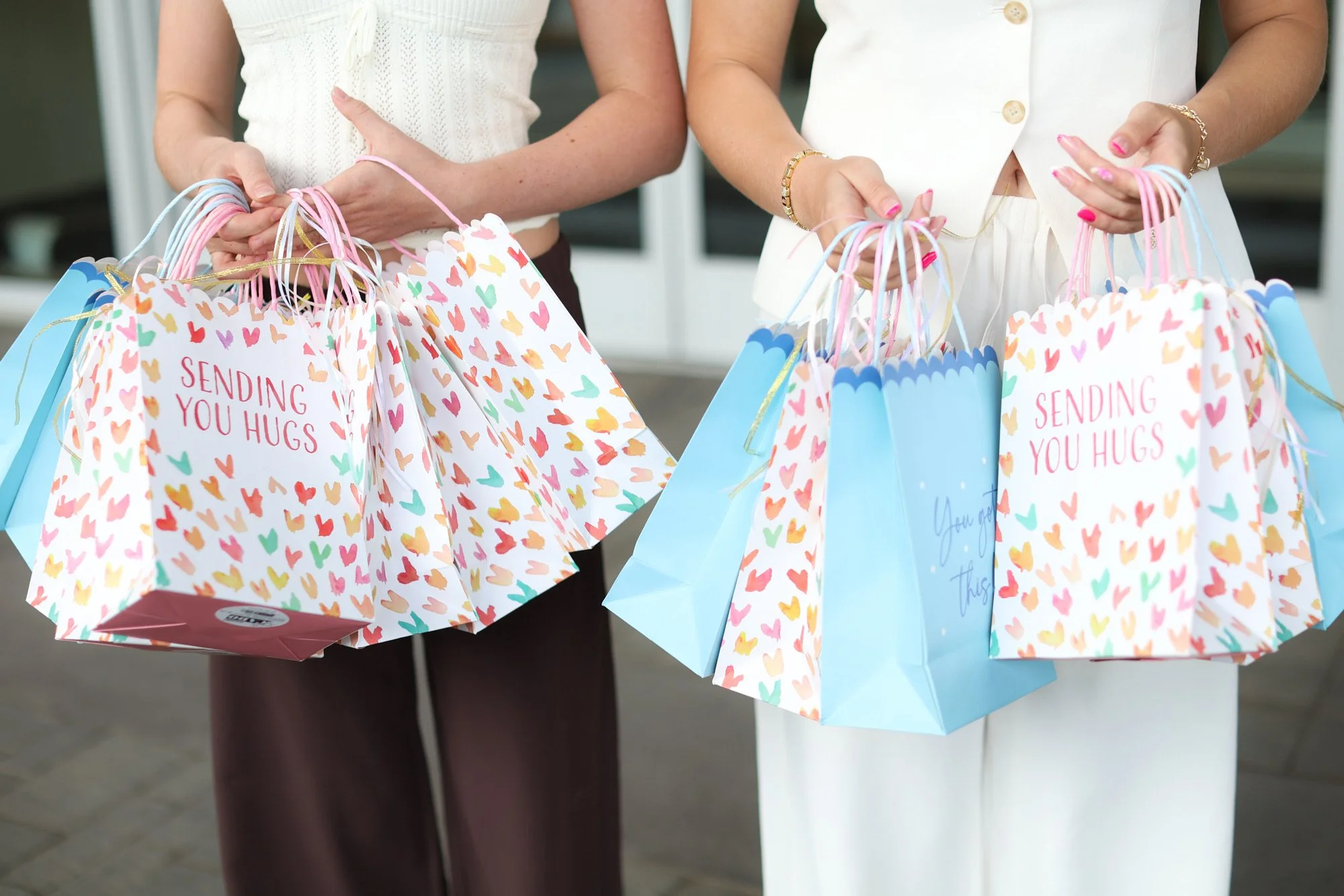 Two women holding colorful gift bags decorated with small hearts and messages, wearing elegant outfits, possibly at a celebration or event.