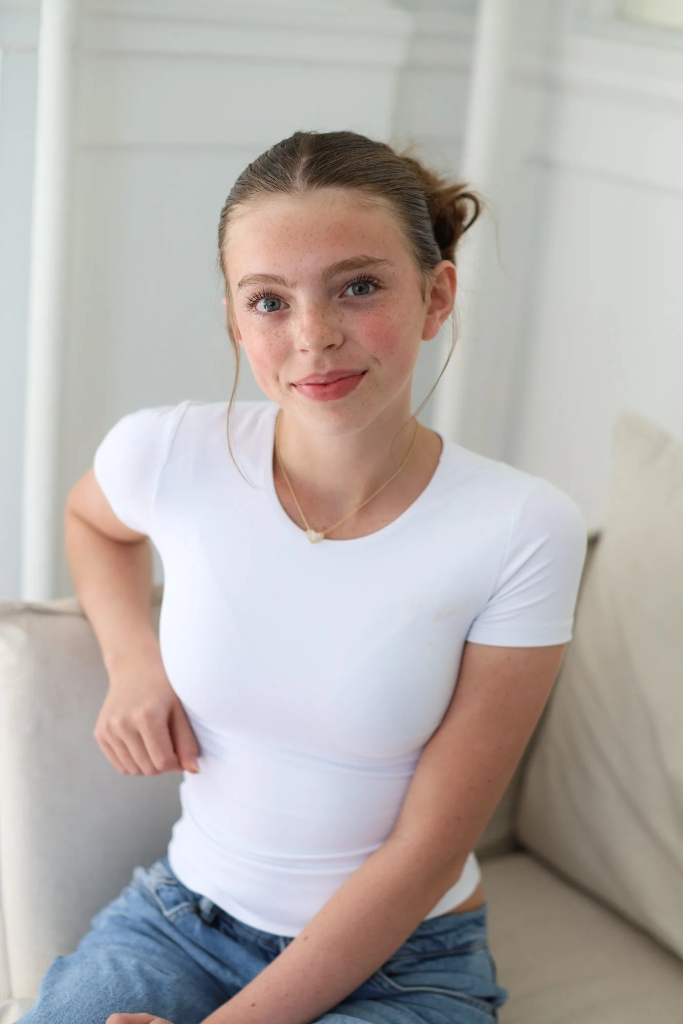 A young woman with brown hair tied back, freckles, and blue eyes, smiling gently while sitting on a light-colored sofa, wearing a white t-shirt and denim jeans, with her arms resting on her lap.