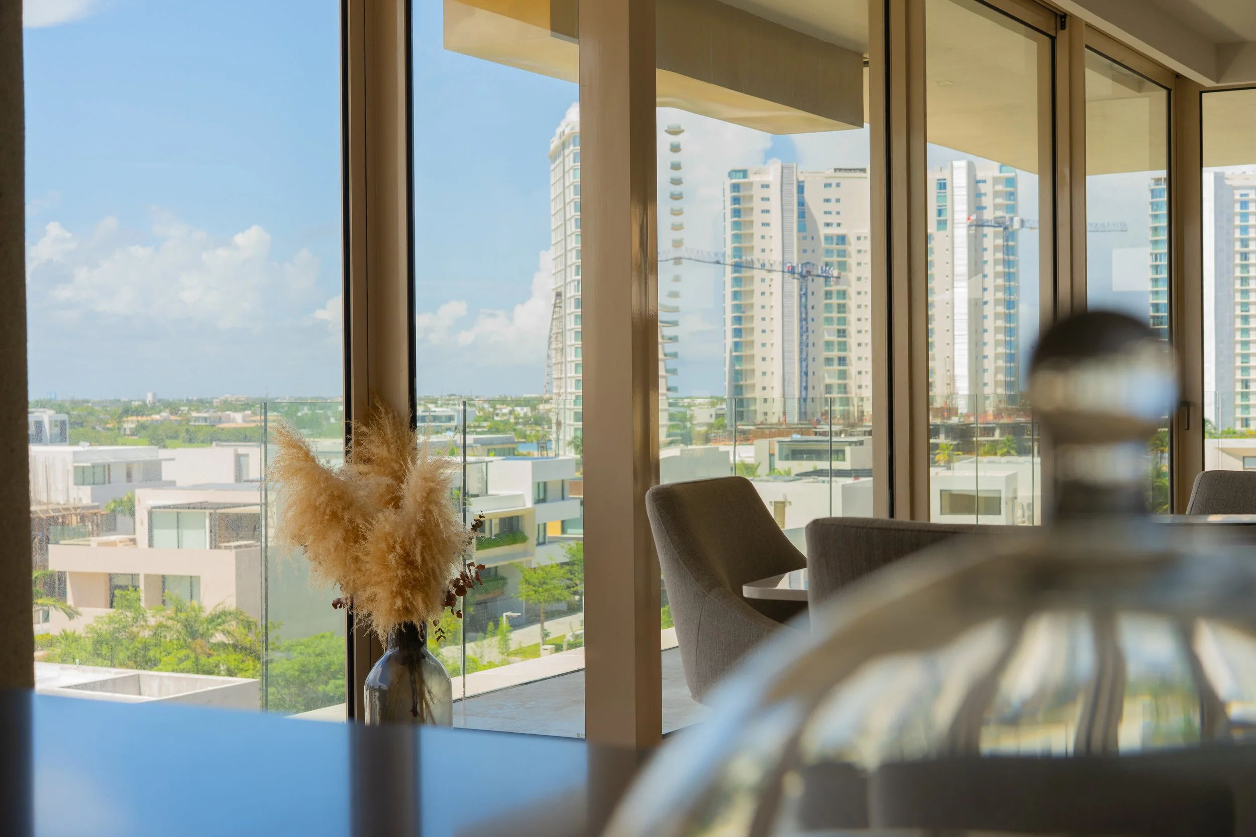 Interior view of a modern high-rise apartment with large floor-to-ceiling windows showcasing a city skyline with tall buildings and cranes. There is a vase with pampas grass on a table in the foreground.