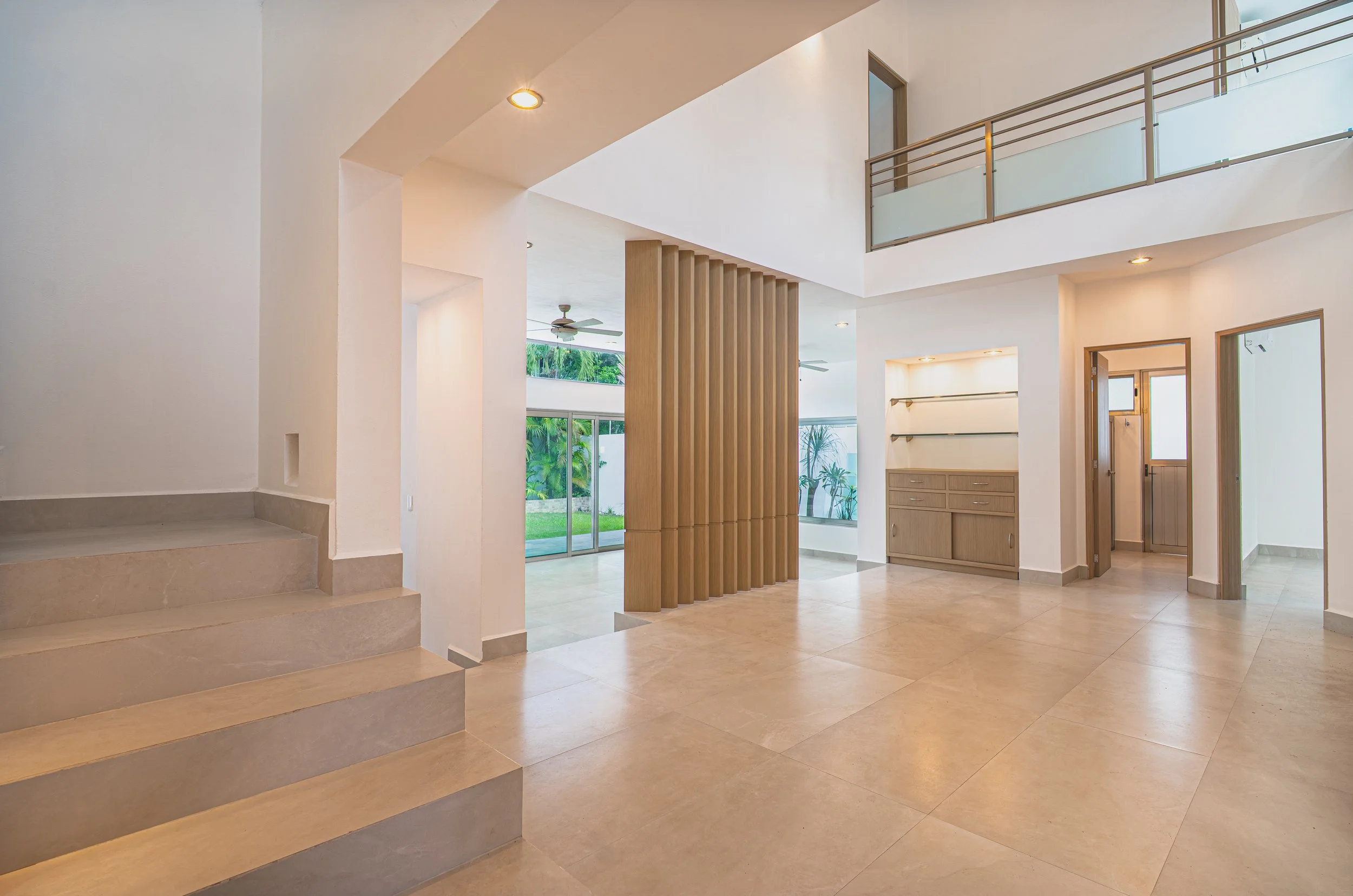 Modern open-concept home interior with tiled floors, floating staircase, wood slat room divider, built-in cabinetry, and glass-railed mezzanine