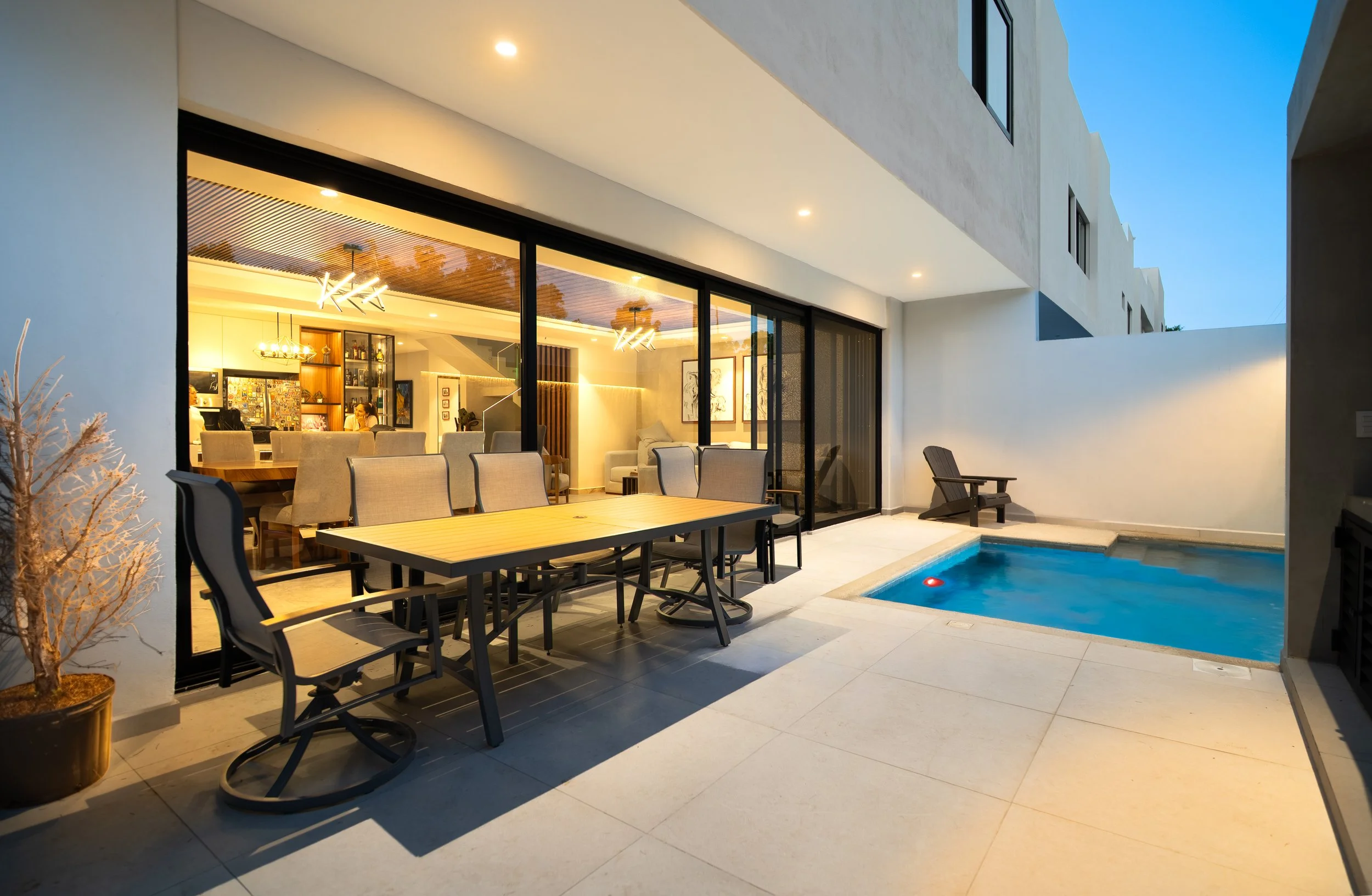 Modern home patio with outdoor dining table, chairs, swimming pool, and glass doors opening to a warmly lit interior.