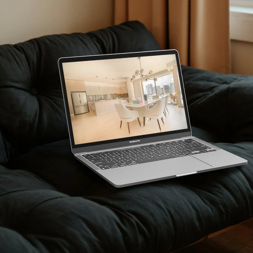 An open MacBook Pro laptop resting on a dark couch, displaying an image of a modern, bright dining and kitchen area with white chairs, a dining table, a kitchen island, and floor-to-ceiling windows showing cityscape views.