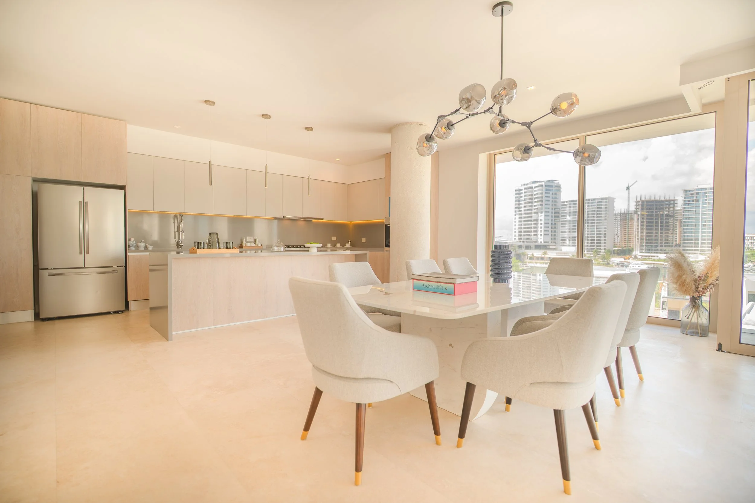 Modern, open-concept kitchen and dining area with large windows, white and light wood furniture, and city skyline view.