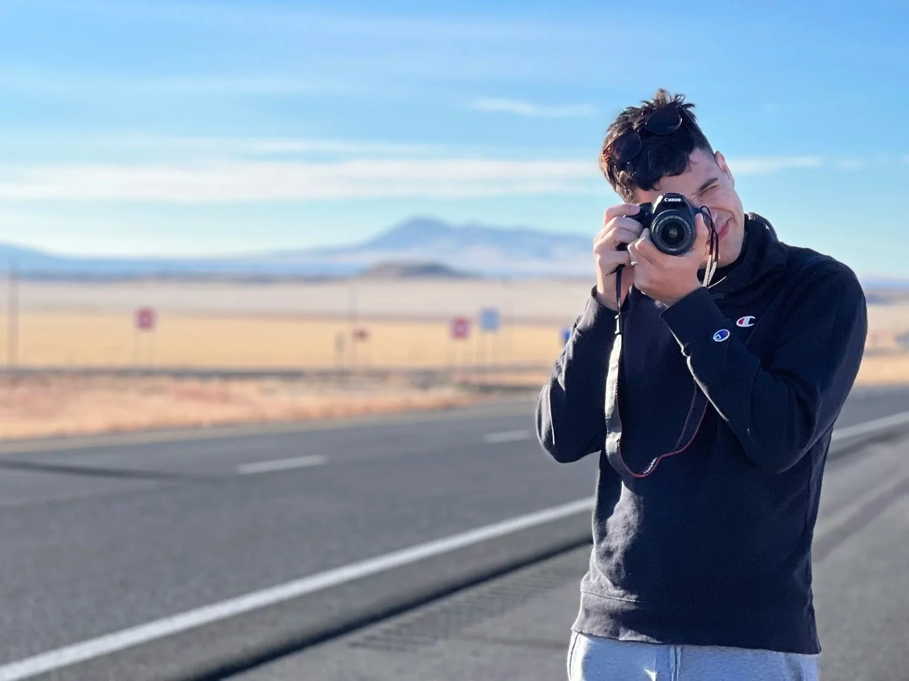 Young man taking a photograph with a camera outdoors on a roadside with open plains and mountains in the distance under a blue sky.