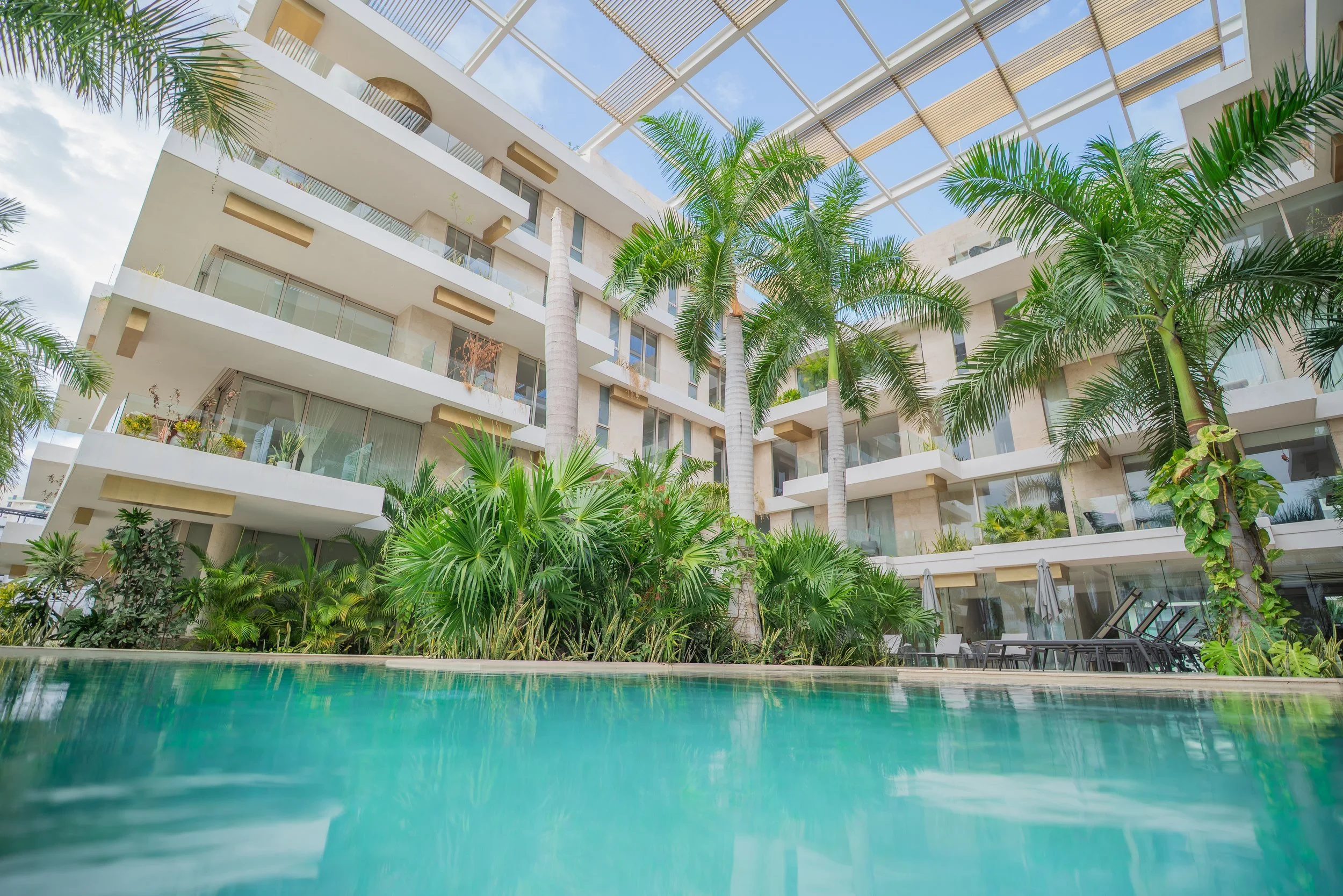 Luxury tropical resort courtyard with turquoise swimming pool, palm trees, lush landscaping, and modern balconies under a pergola roof