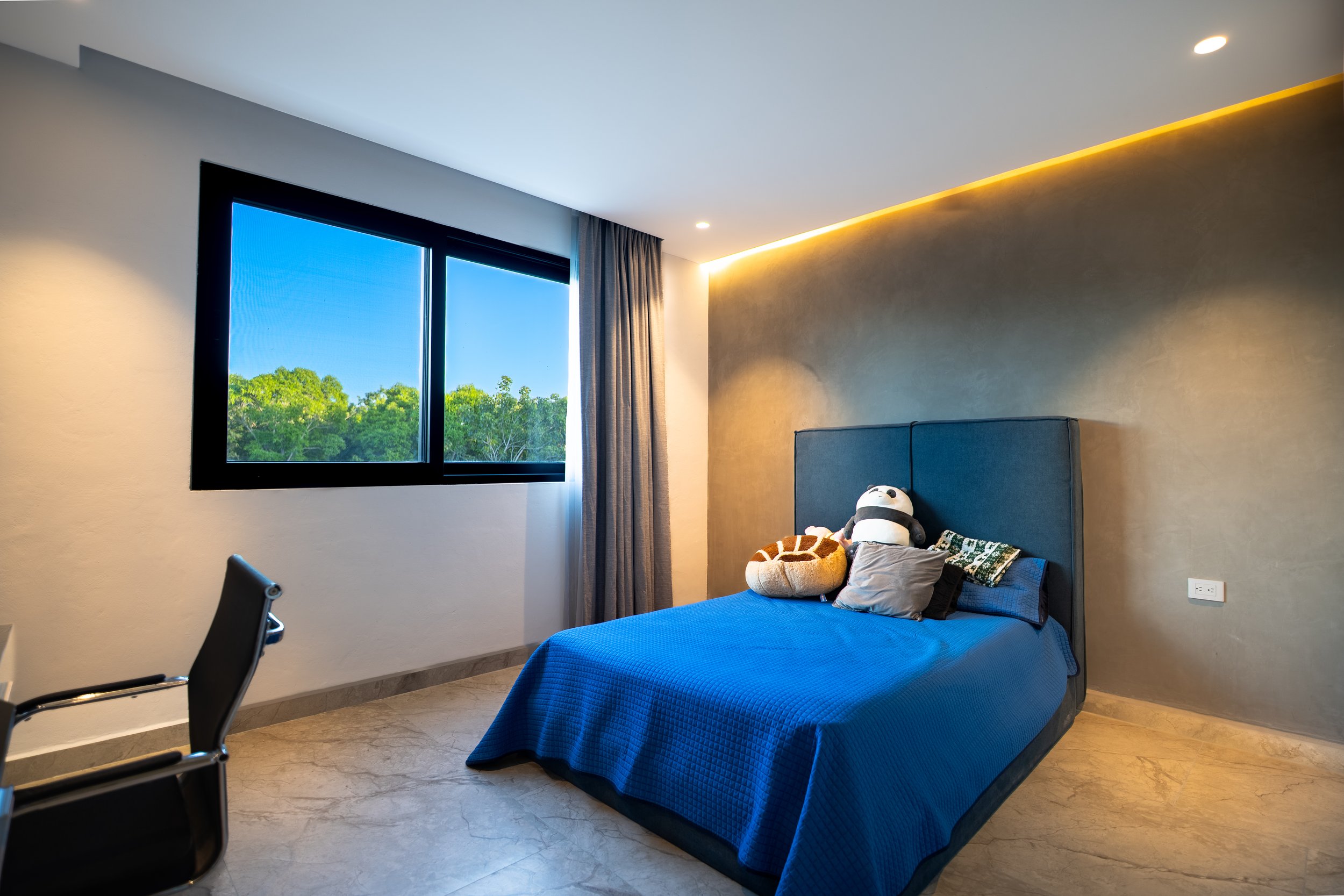 Modern bedroom with a blue quilted bed, plush toys, large window, and gray accent wall with LED lighting