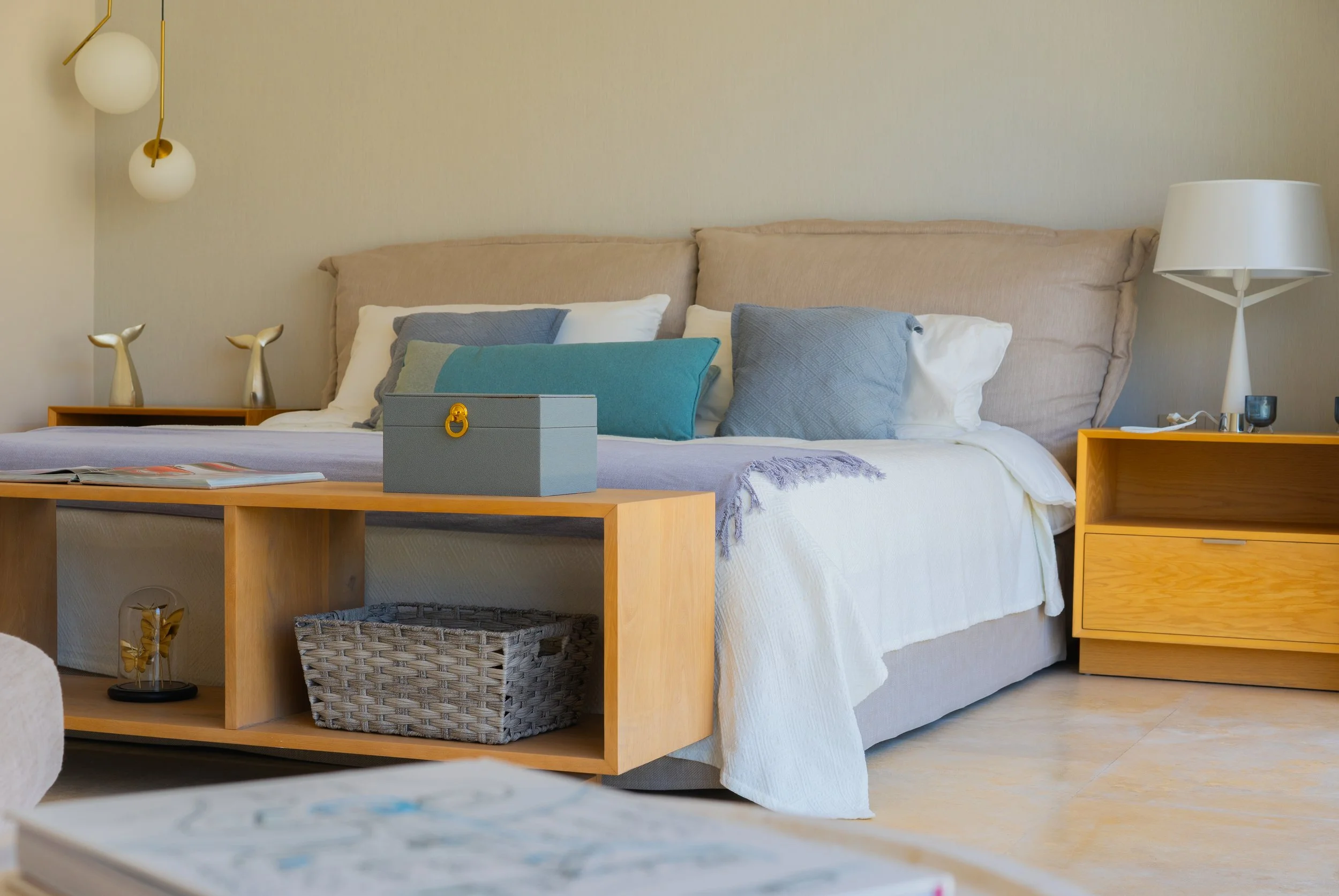 Modern bedroom with a neatly made bed, blue and beige pillows, wooden bench, storage basket, bedside table, and contemporary lamps