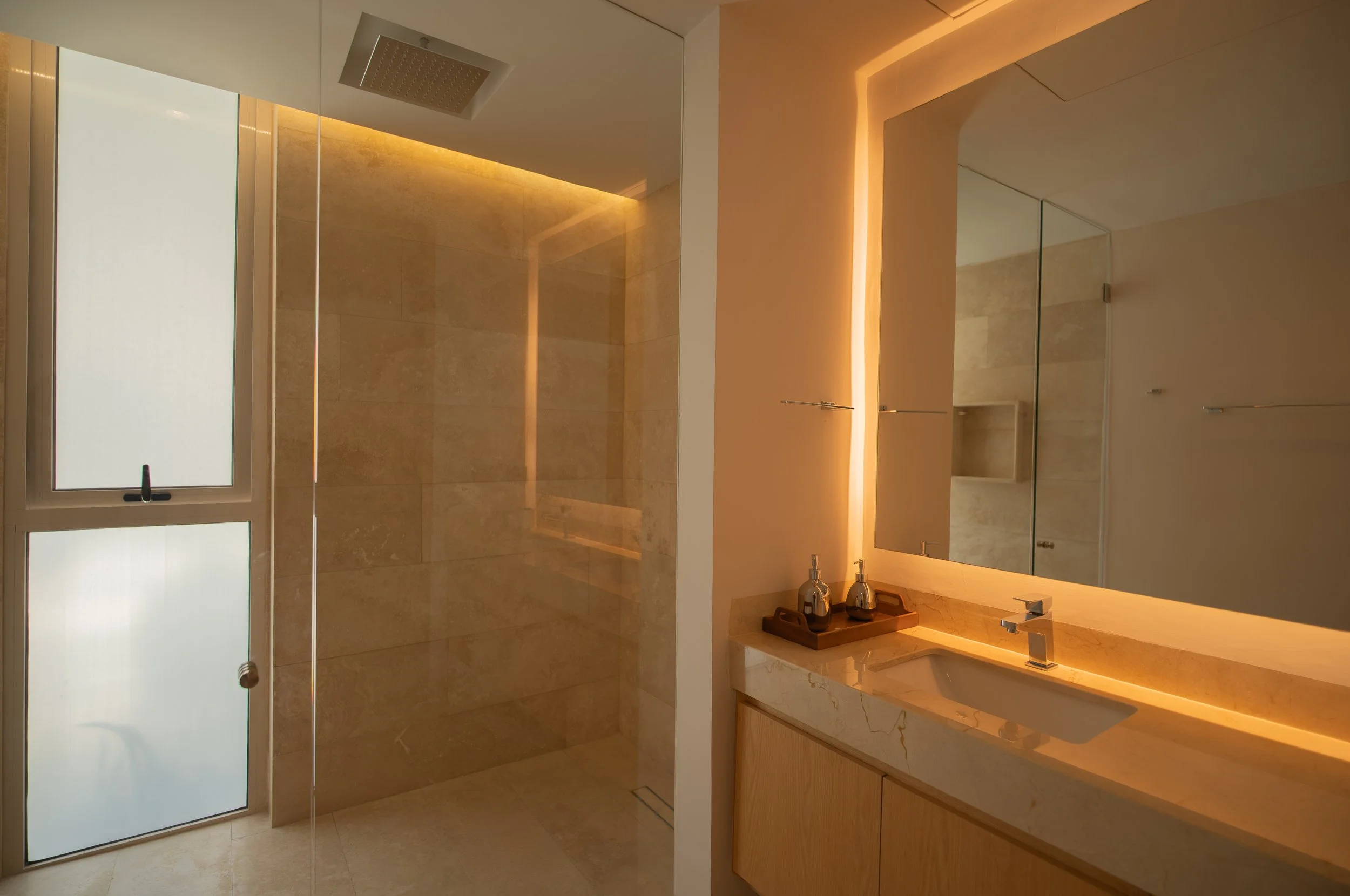 Modern luxury bathroom with glass-enclosed rain shower, marble vanity, large mirror, and warm accent lighting