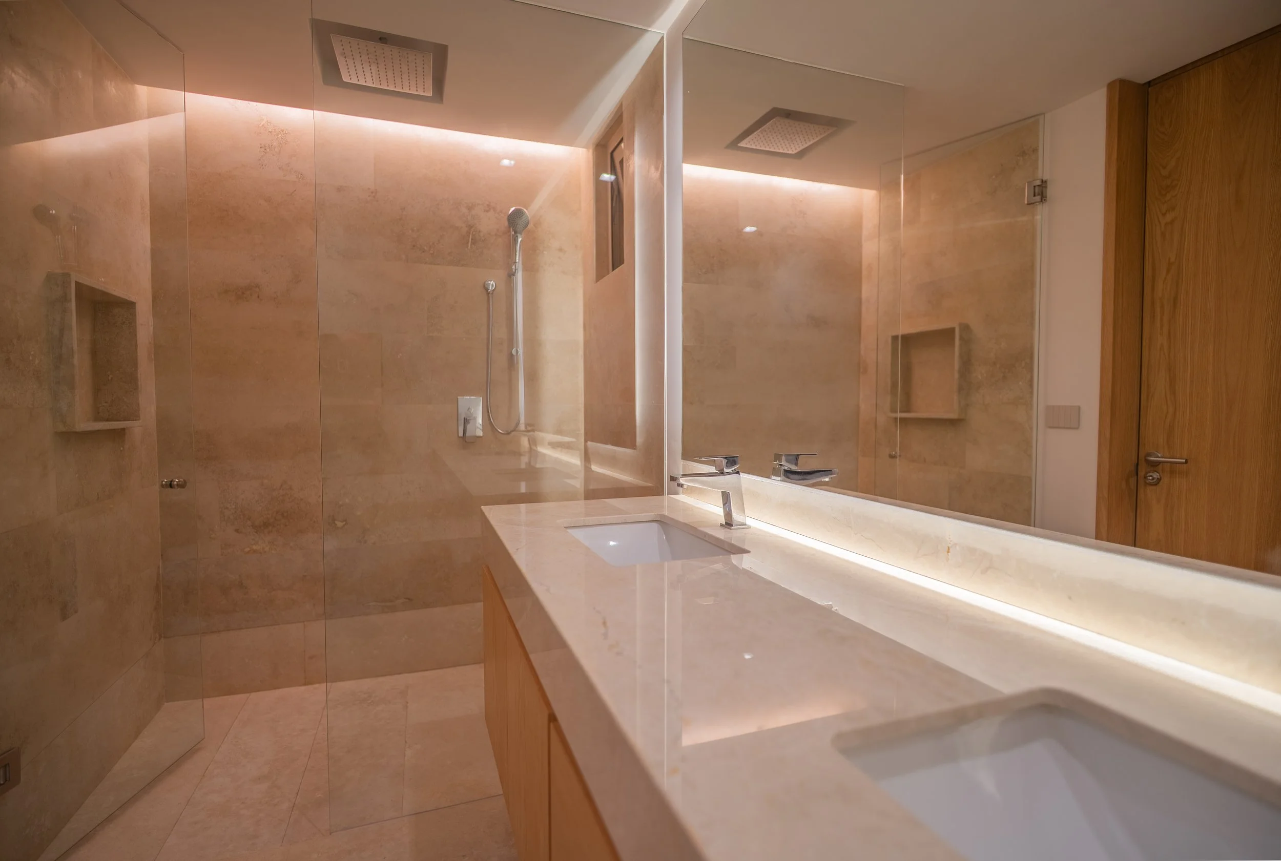 Modern bathroom with beige marble tiles, a large mirror, a double sink vanity with a marble countertop, a wall-mounted shower with a handheld showerhead, and a ceiling-mounted rainfall showerhead.