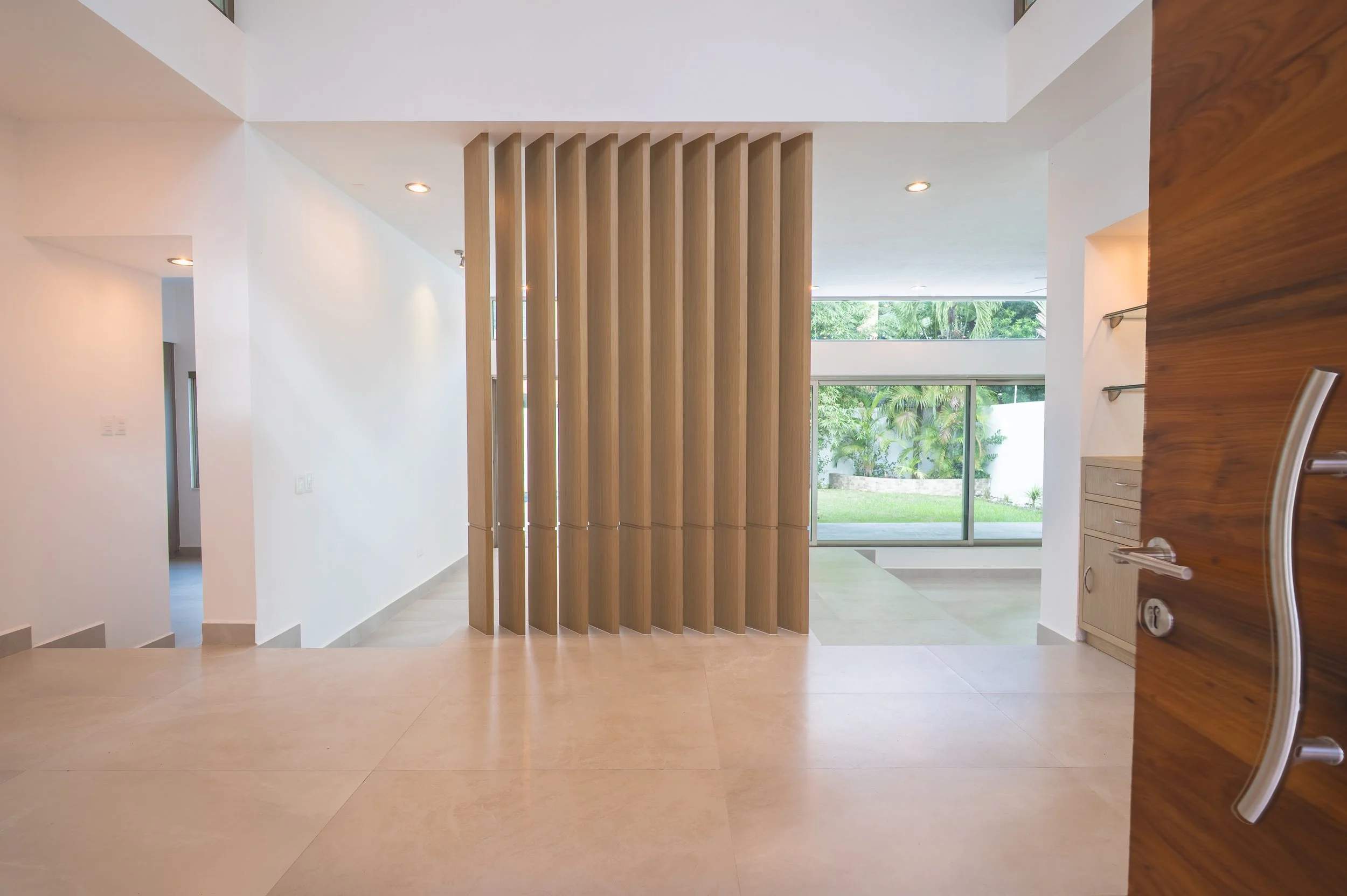 Interior view of a modern house with white walls, a wooden room divider, large window/views of greenery, and tiled flooring.