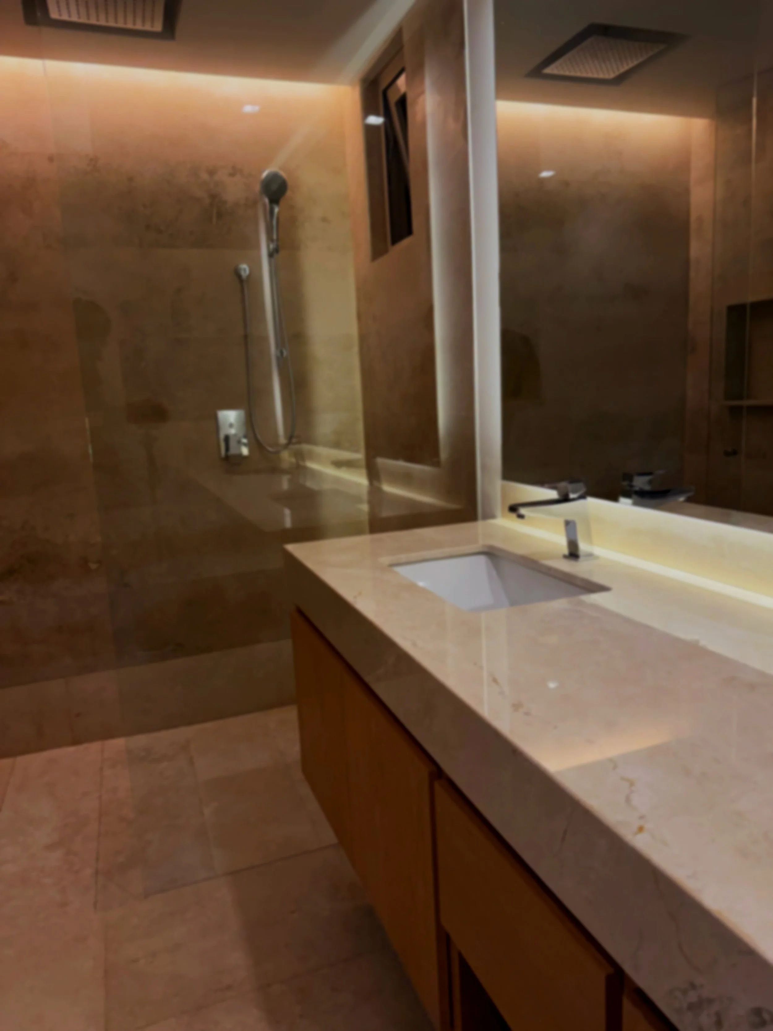 Bathroom with beige marble countertop, undermount sink, wooden cabinets, wall with a showerhead, mirror, and multiple ventilation panels on ceiling.