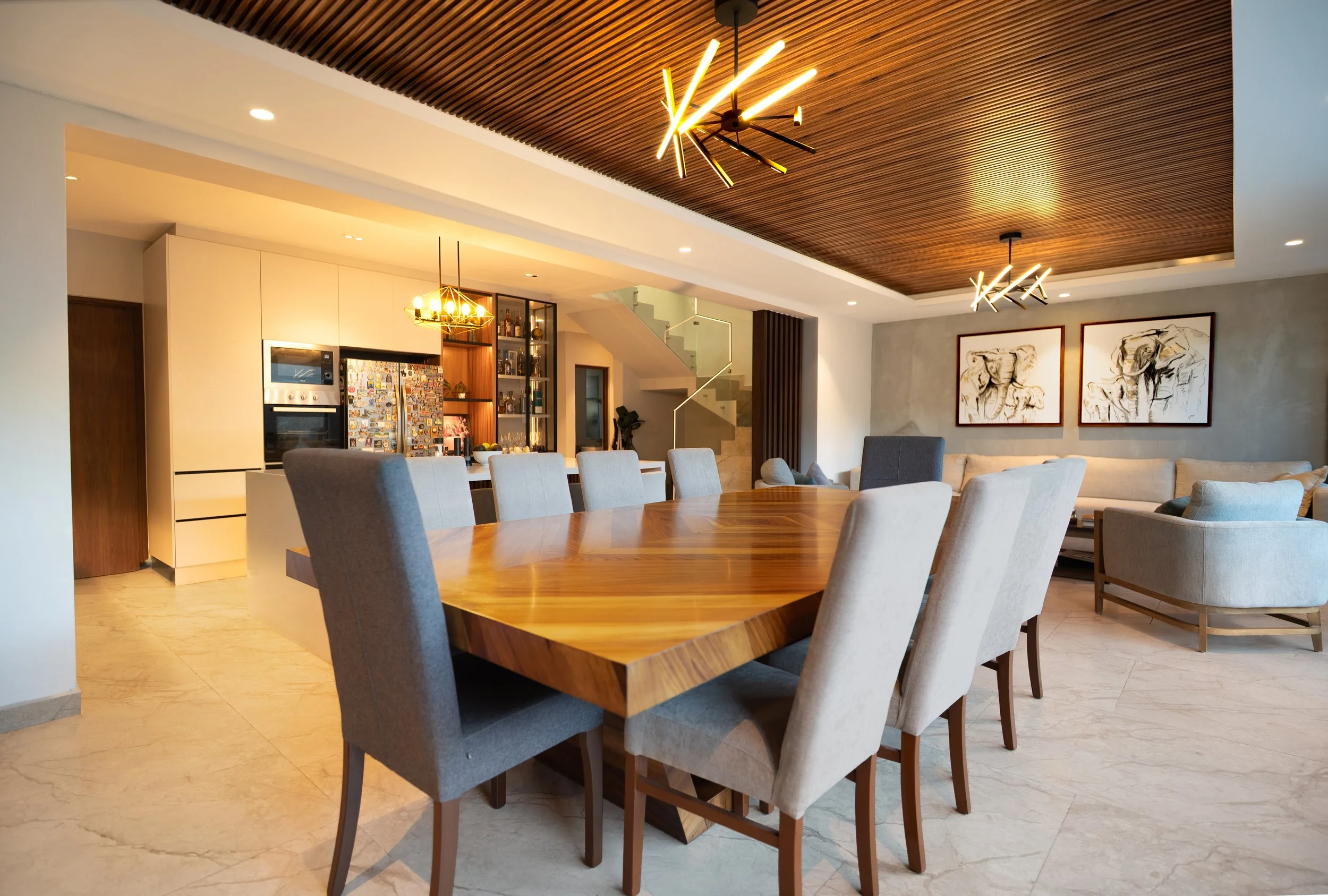 Modern open-concept dining and living room with a wooden table, upholstered chairs, contemporary lighting, kitchen, staircase, and framed artwork