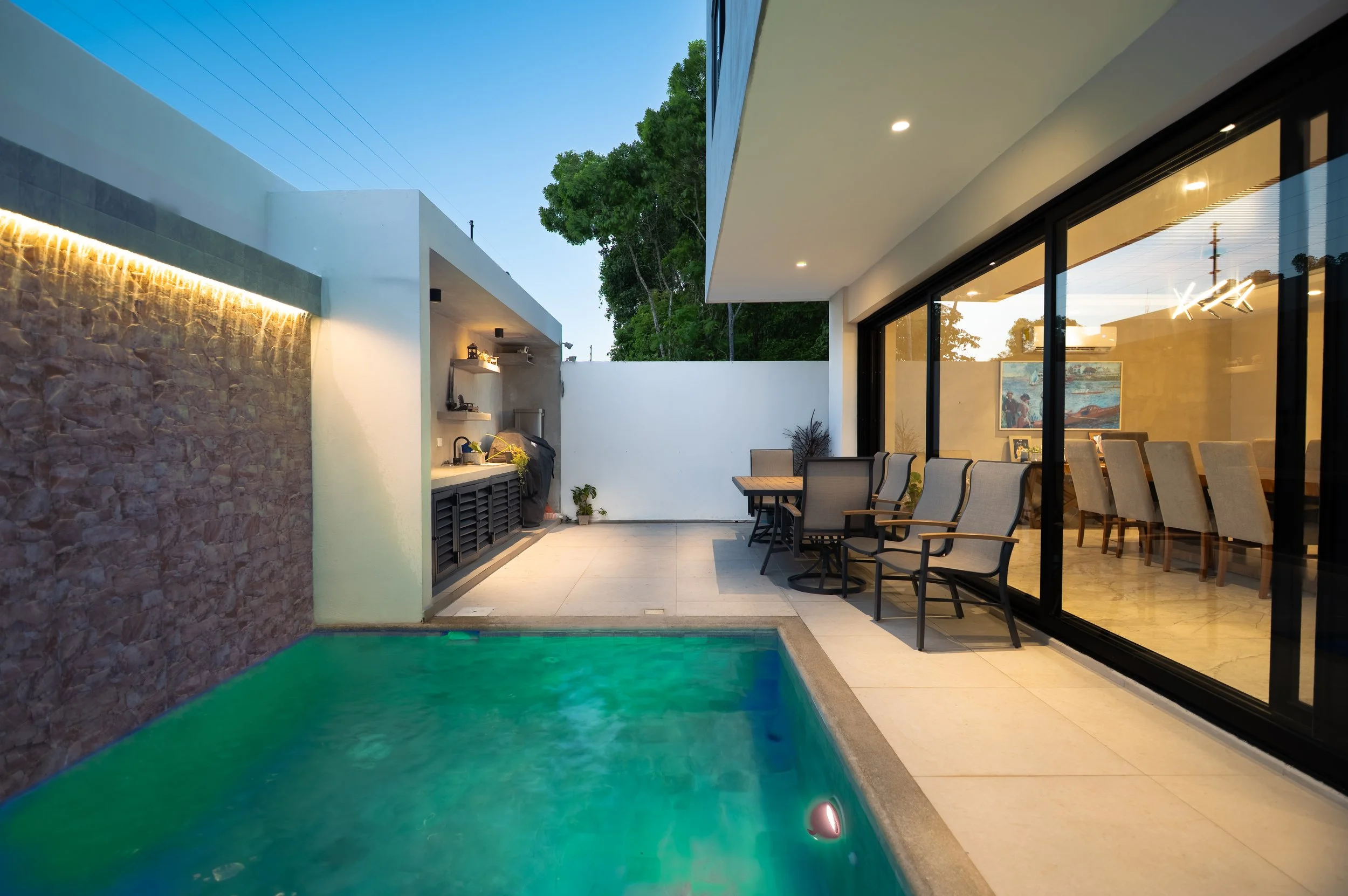 Modern backyard patio with a swimming pool, outdoor kitchen, dining area, and glass-walled living room at dusk