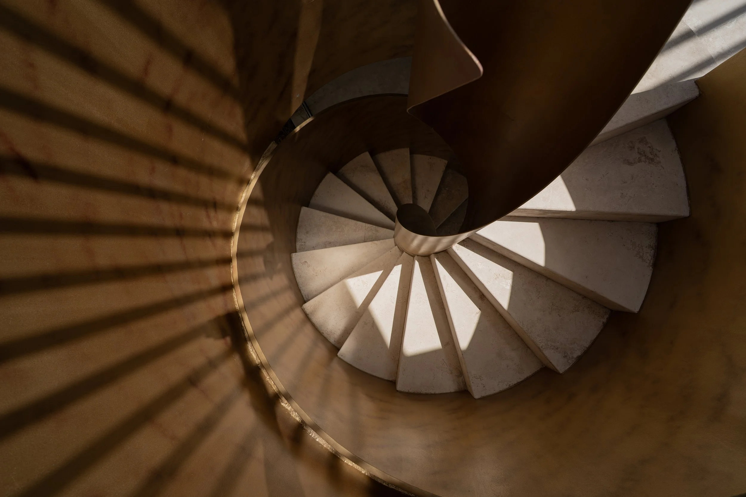 Top-down view of a spiral staircase with concrete steps, a central metal pole, and dramatic sunlight and shadows.