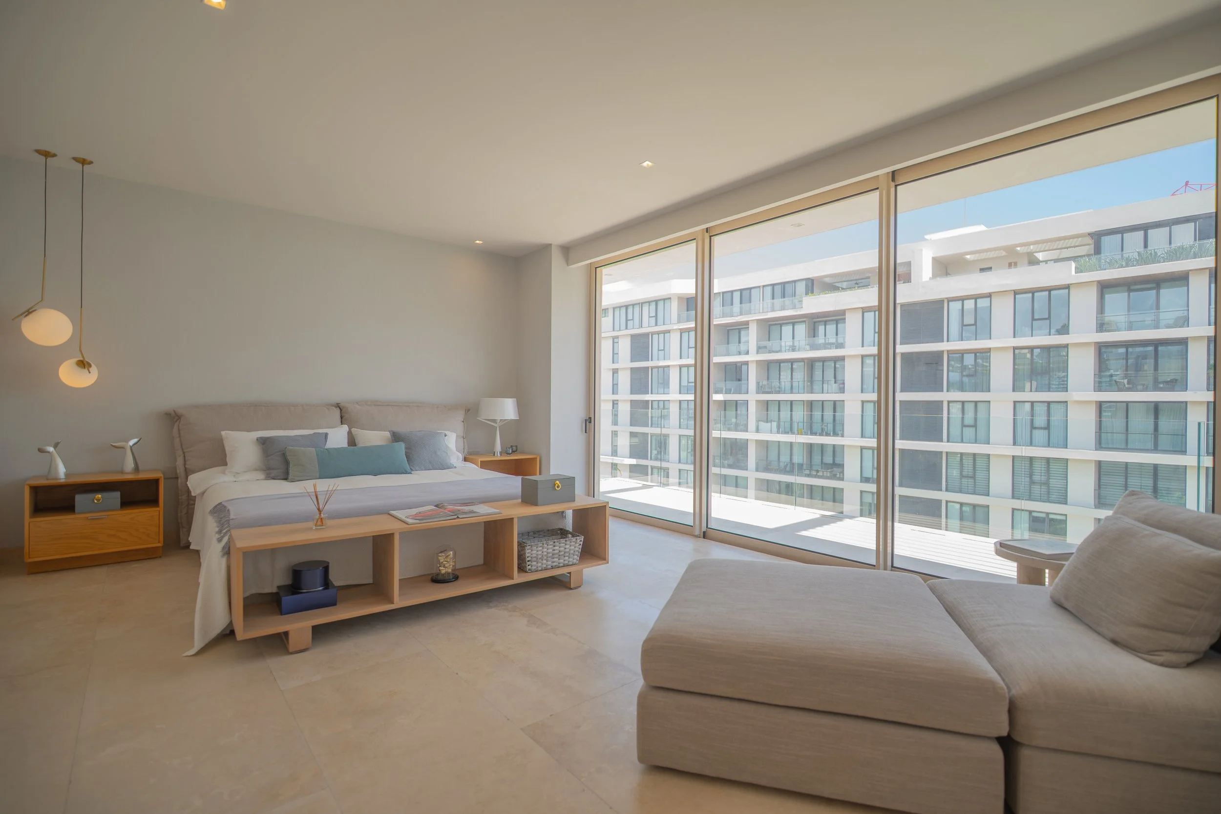 Modern apartment bedroom with bed, sectional sofa, contemporary furnishings, and floor-to-ceiling windows overlooking a neighboring building