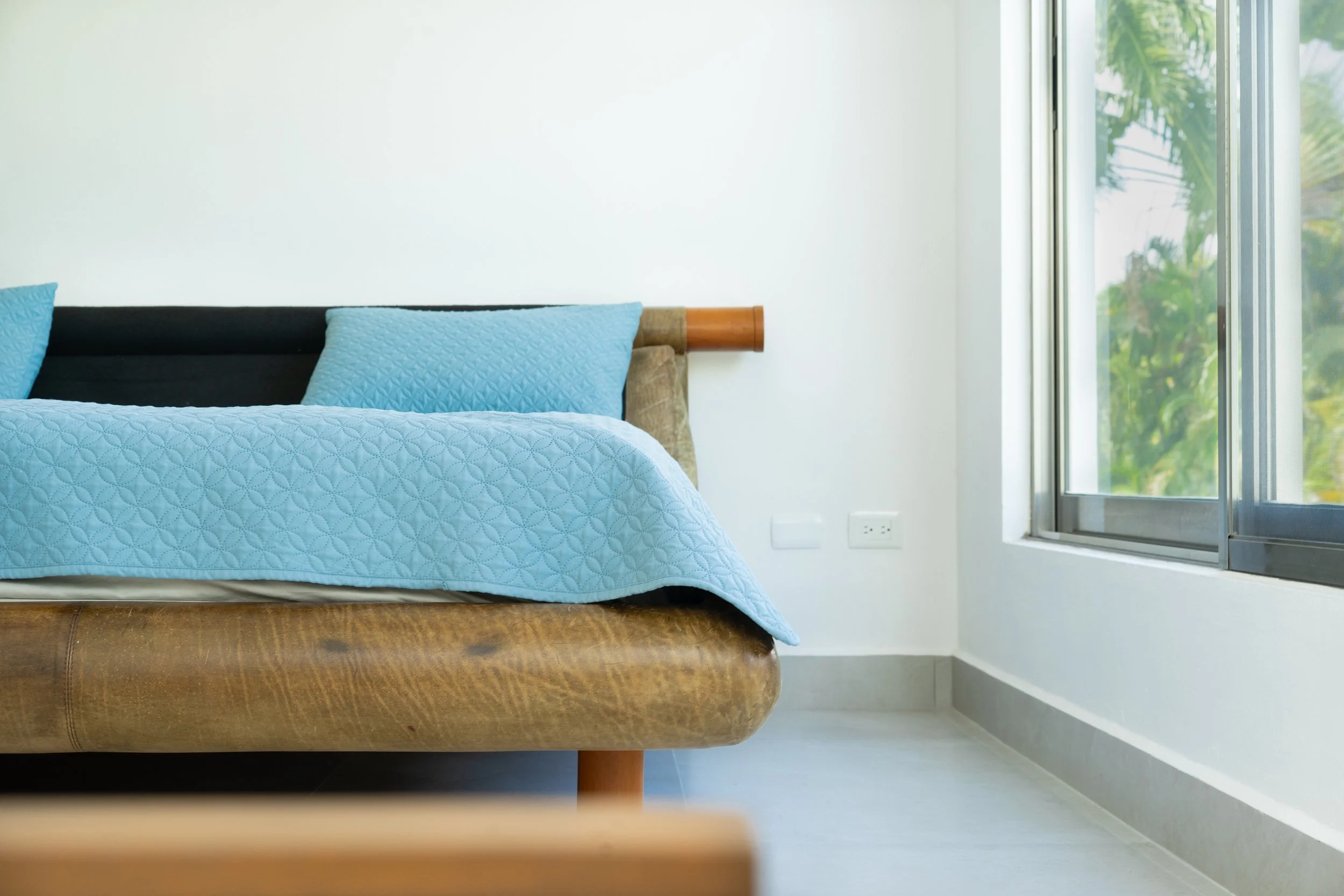 A close-up of a bed with a brown leather headboard and a blue quilted bedspread, beside a large window with a view of green foliage.