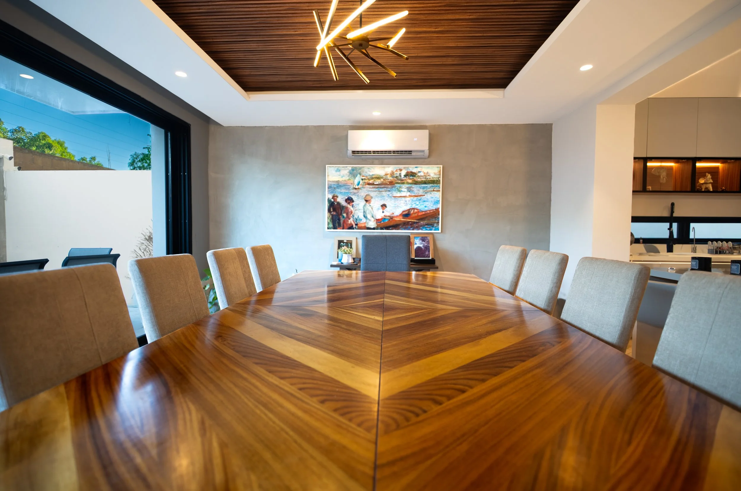 Modern dining room with a large wood dining table, upholstered chairs, geometric pendant light, coastal painting, and adjoining kitchen.