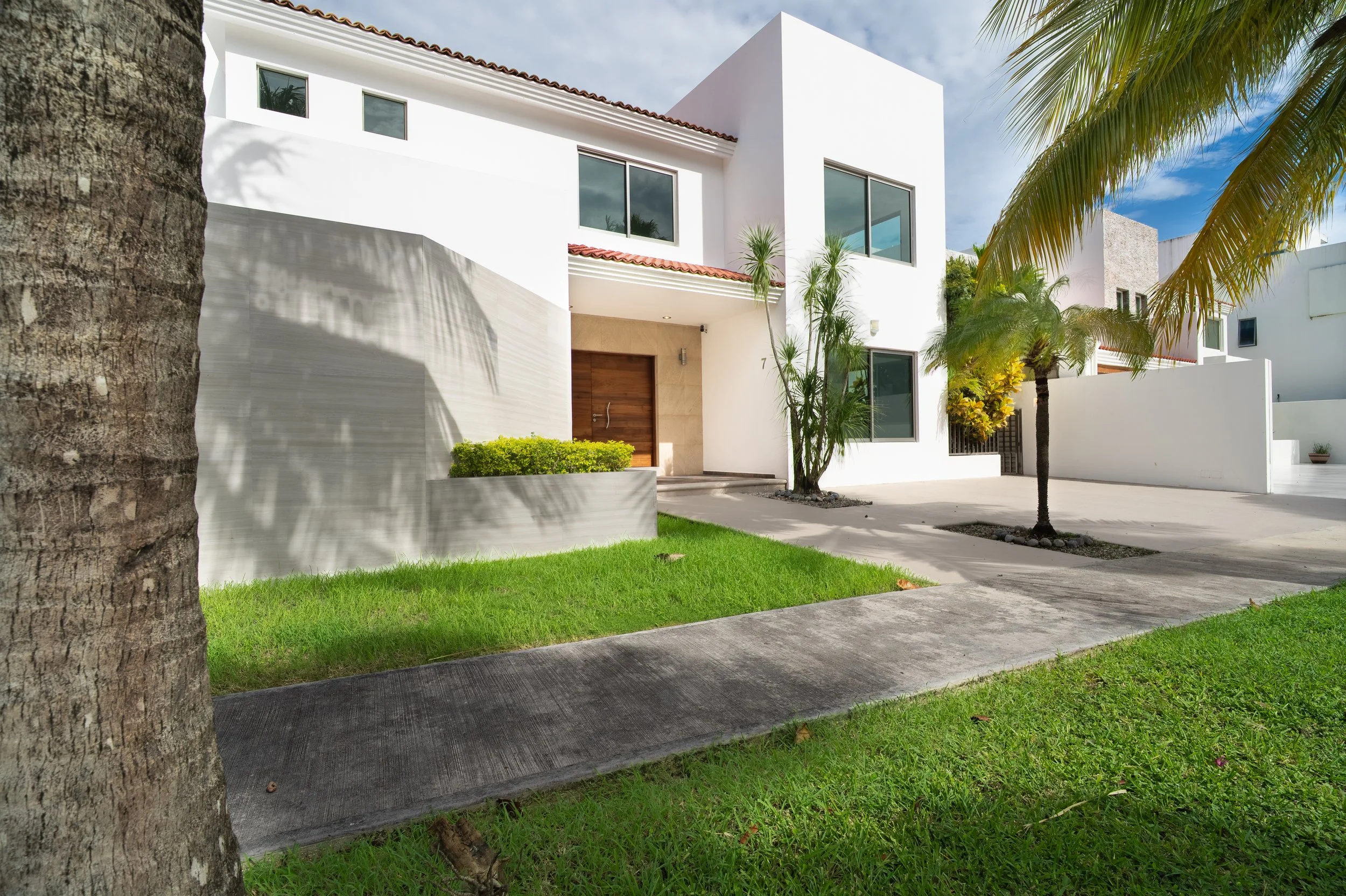 Modern white two-story house with red tile roof, tropical palm trees, landscaped lawn, and driveway