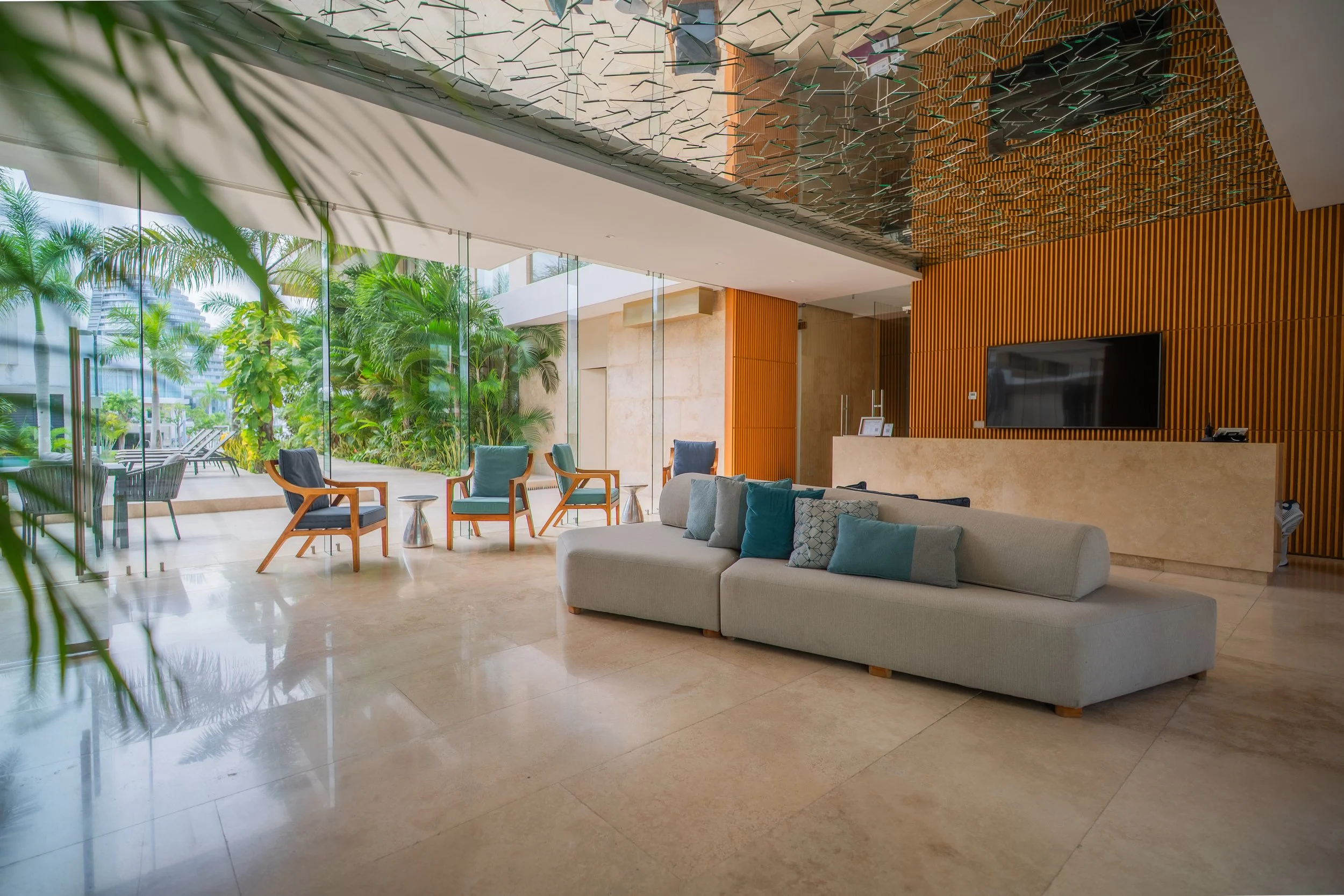 Modern hotel lobby with gray sofas, lounge chairs, tropical landscaping, wood-paneled walls, and a reception desk