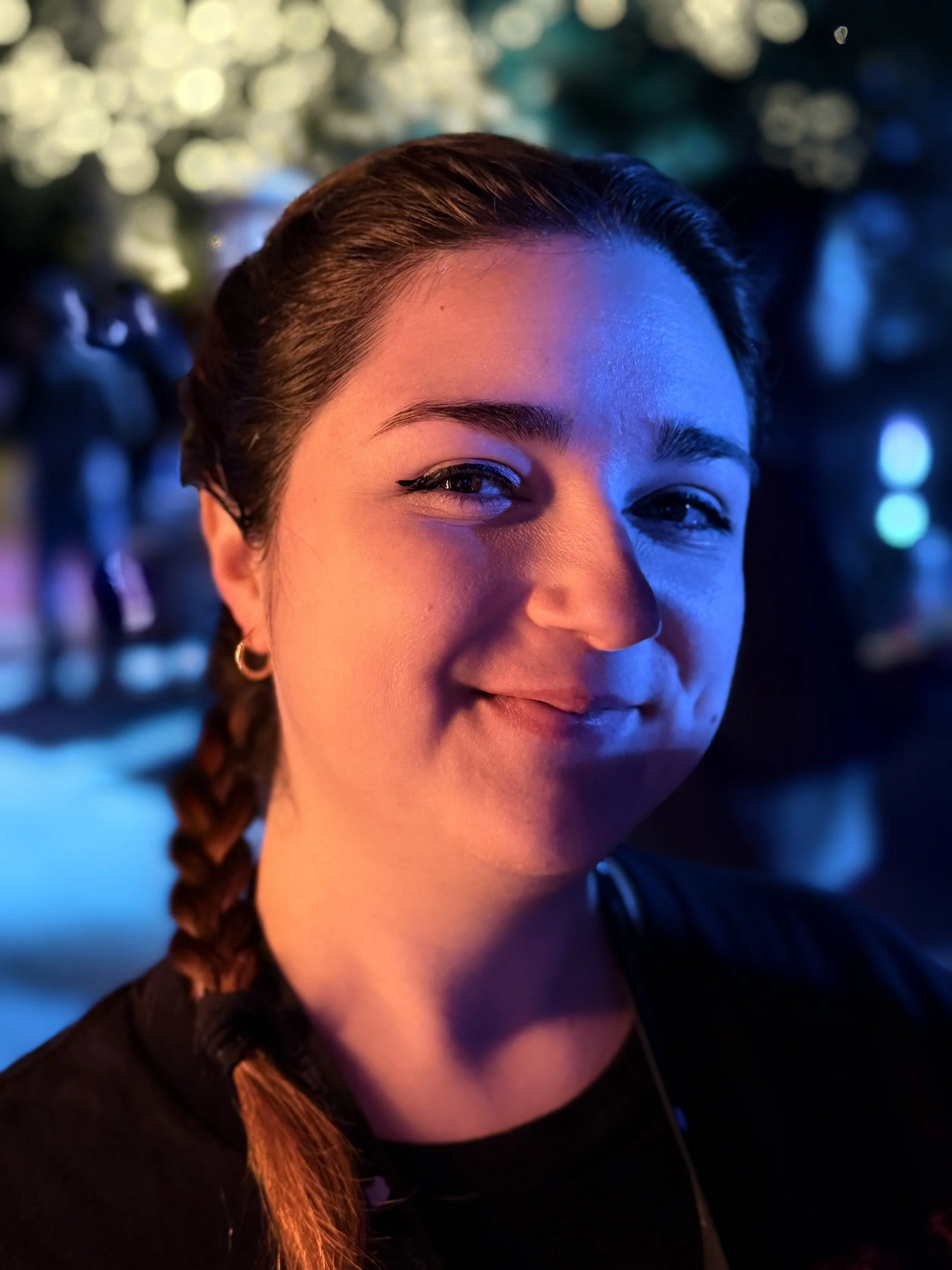 Close-up of a young woman smiling with braided hair, night setting with colorful bokeh lights in the background.
