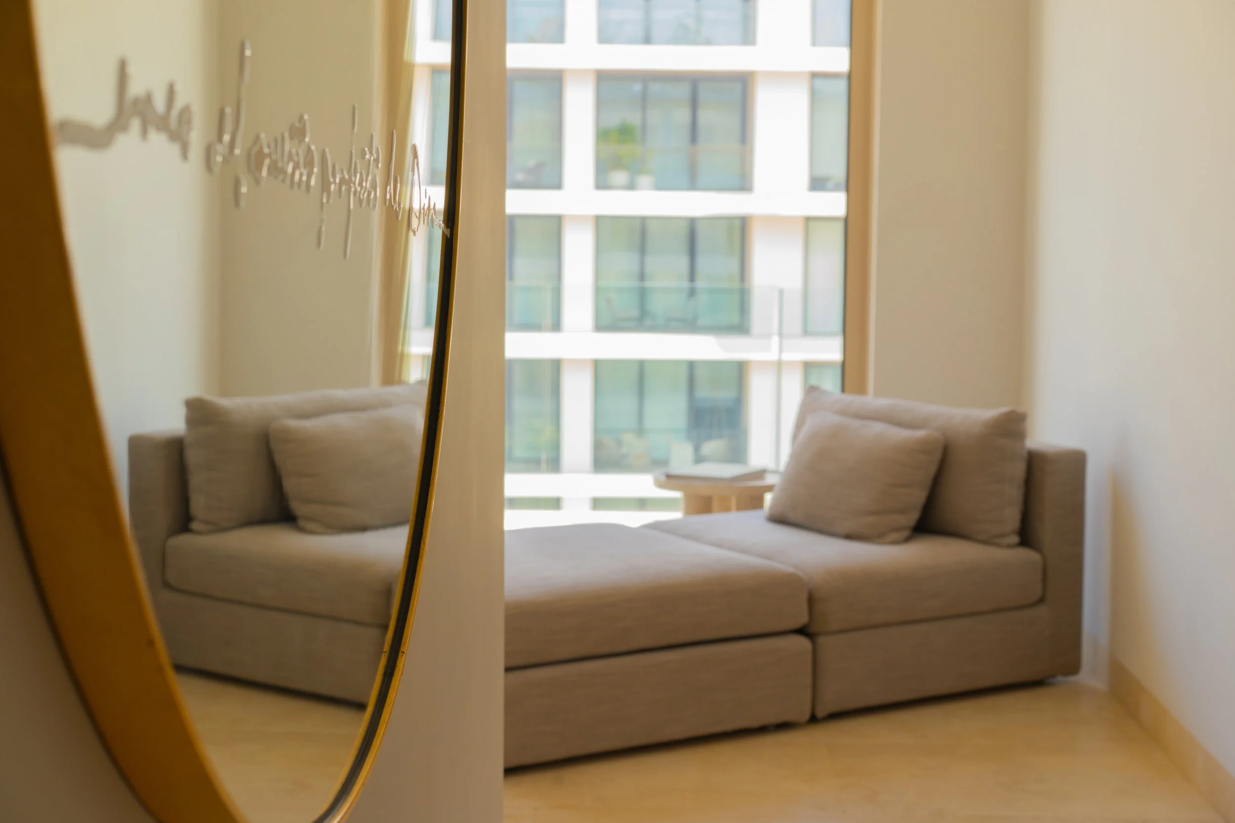 A mirror reflecting a beige sofa with matching pillows and a wooden side table in front of large picture windows in a modern apartment.
